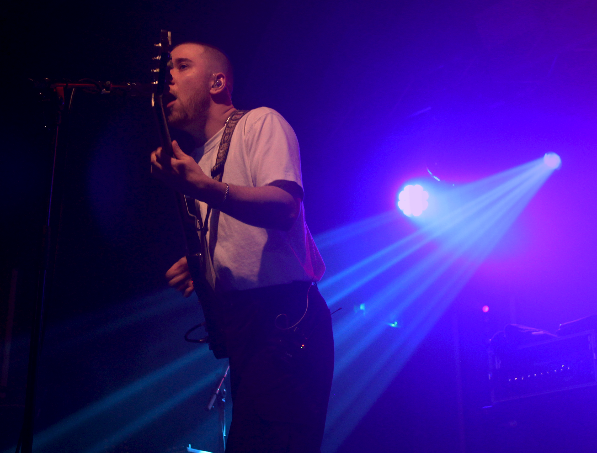 A male musician with a shaved head and beard singing into a microphone while playing an electric guitar on stage, illuminated by blue and purple stage lights.