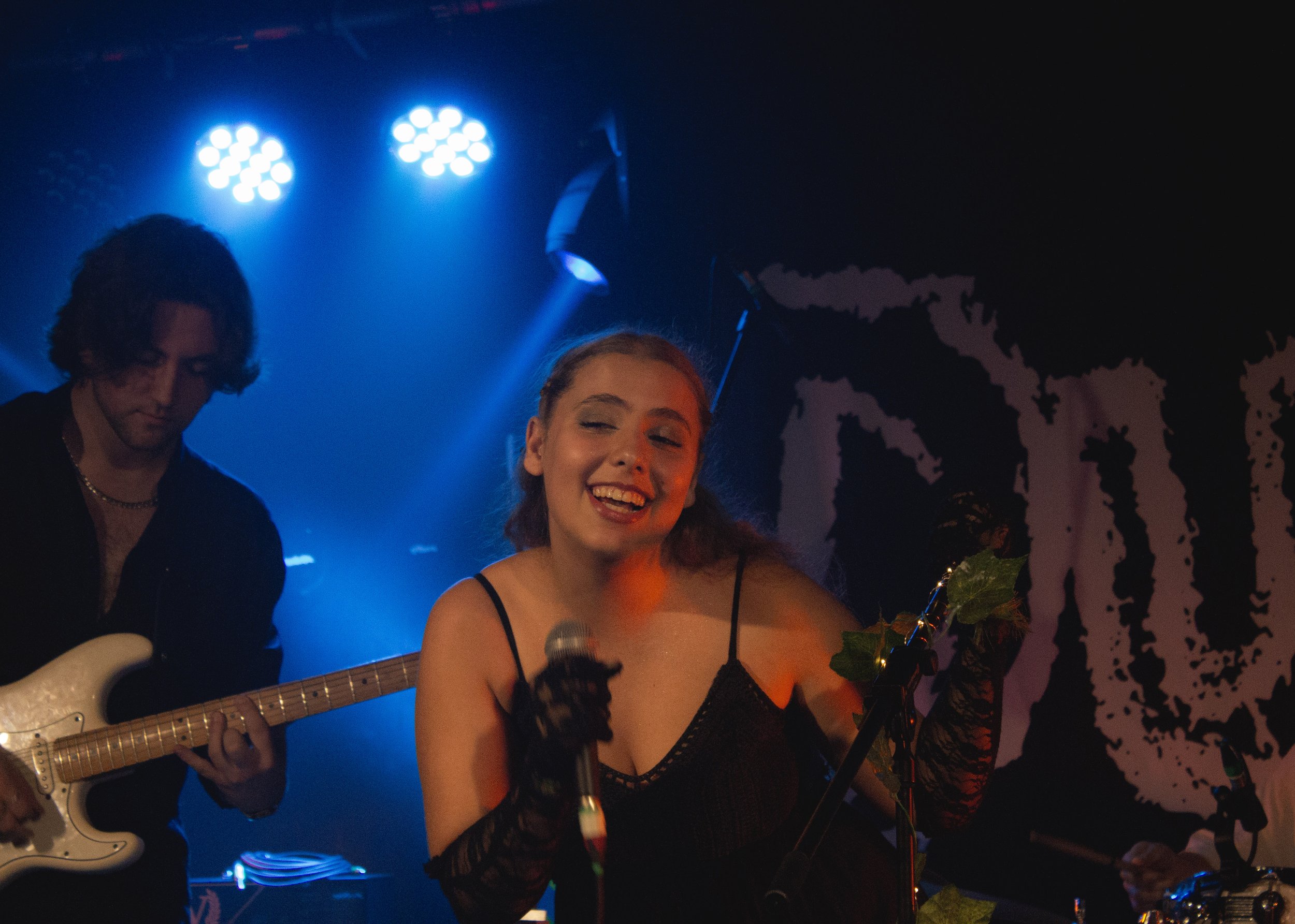 A woman singing into a microphone on stage, smiling, wearing a black dress and black lace gloves, with a guitarist playing in the background under blue stage lighting.