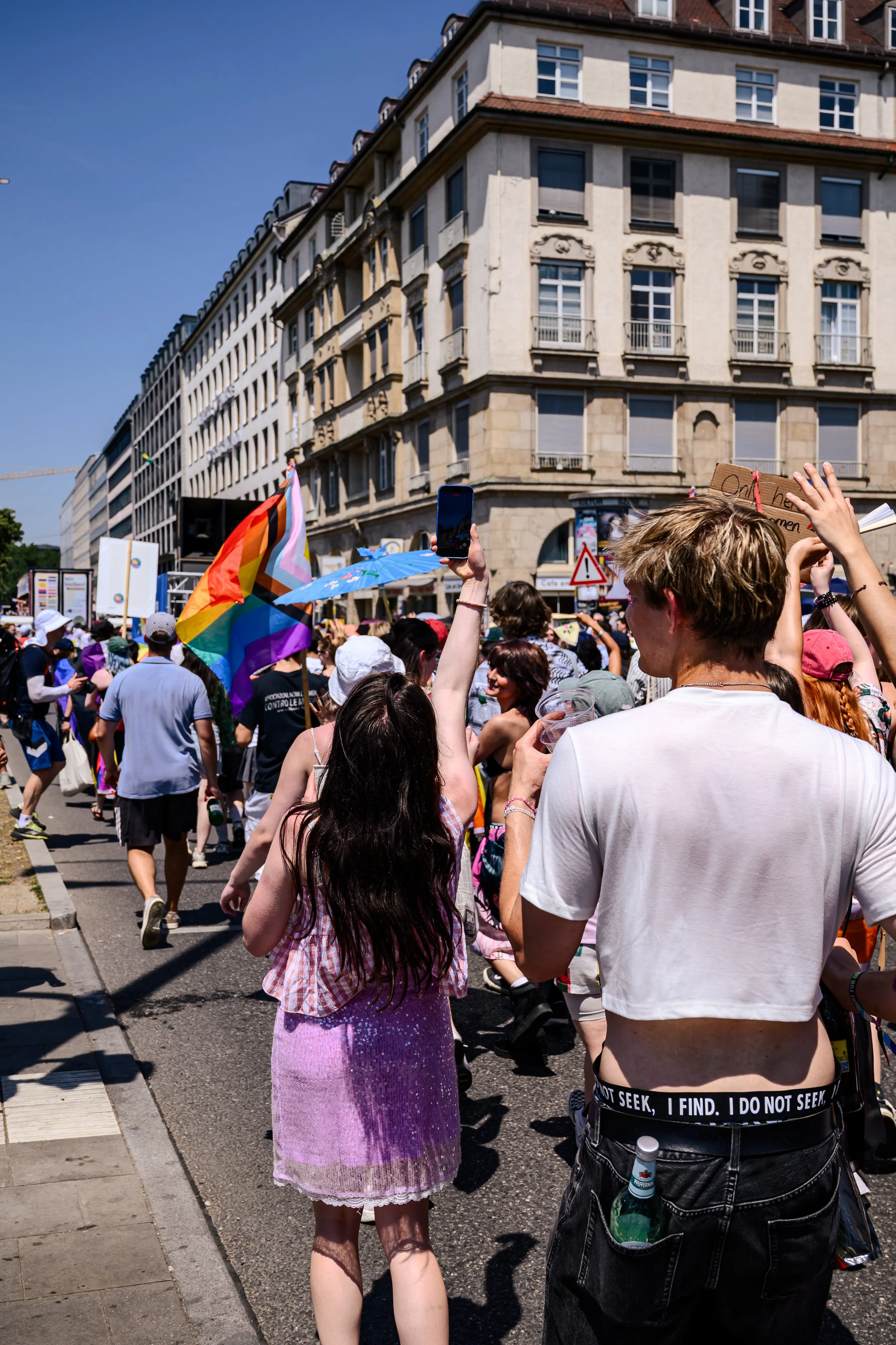 Menschen nehmen an einer Demonstration teil, jemand hält eine Regenbogenfahne, in der Hand eines Mannes ist eine Bierflasche in der Tasche. Menschen stehen auf einer Stadtstraße, im Hintergrund sind Gebäude zu sehen.