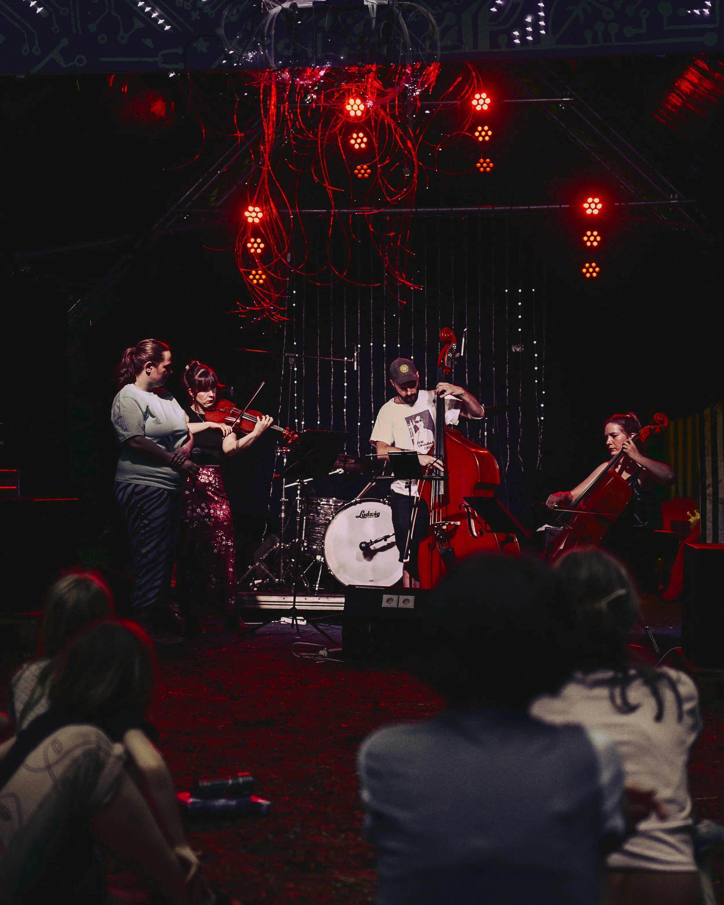 Musiker spielen auf einer Bühne bei einem Konzert, im Hintergrund leuchtende rote Lichter und eine Wand mit technologischen Mustern, im Vordergrund sind Zuschauer zu sehen.