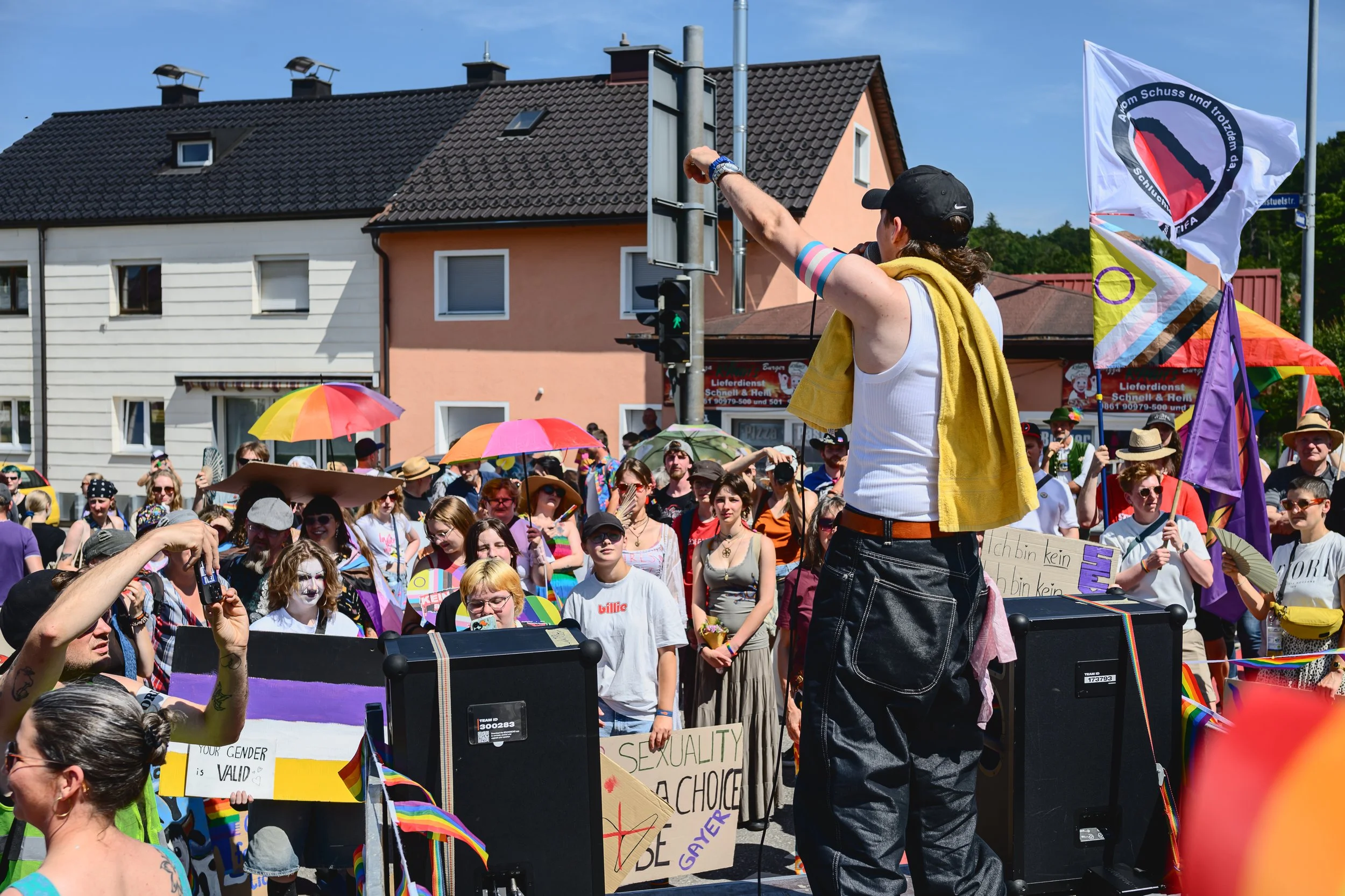 Menschen bei einer Pride-Parade, tragen bunte Kleidung und halten Schilder mit Nachrichten für Gleichstellung und Akzeptanz. Ein Anführer steht auf einem Podest und winkt in die Menge. Es sind Flaggen und Regenschirme mit Regenbogenfarben zu sehen.