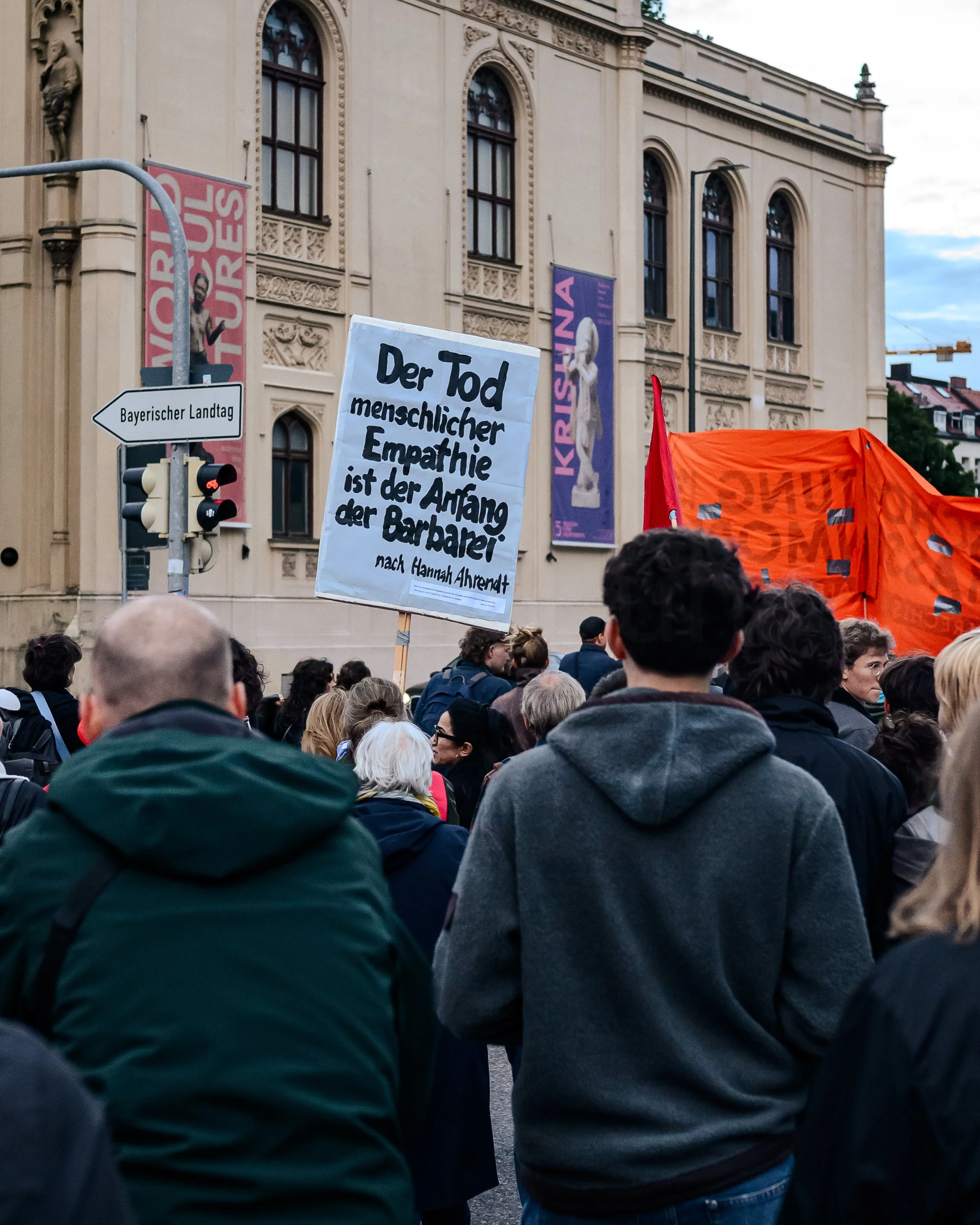 Menschen versammeln sich bei einer Demonstration, einige tragen Schilder mit Botschaften. Im Hintergrund ein historisches Gebäude mit Fenstern und Fassadendekoration, zwei vertikale Banner mit Aufschriften, eine Straßenlaterne und eine Ampel. 