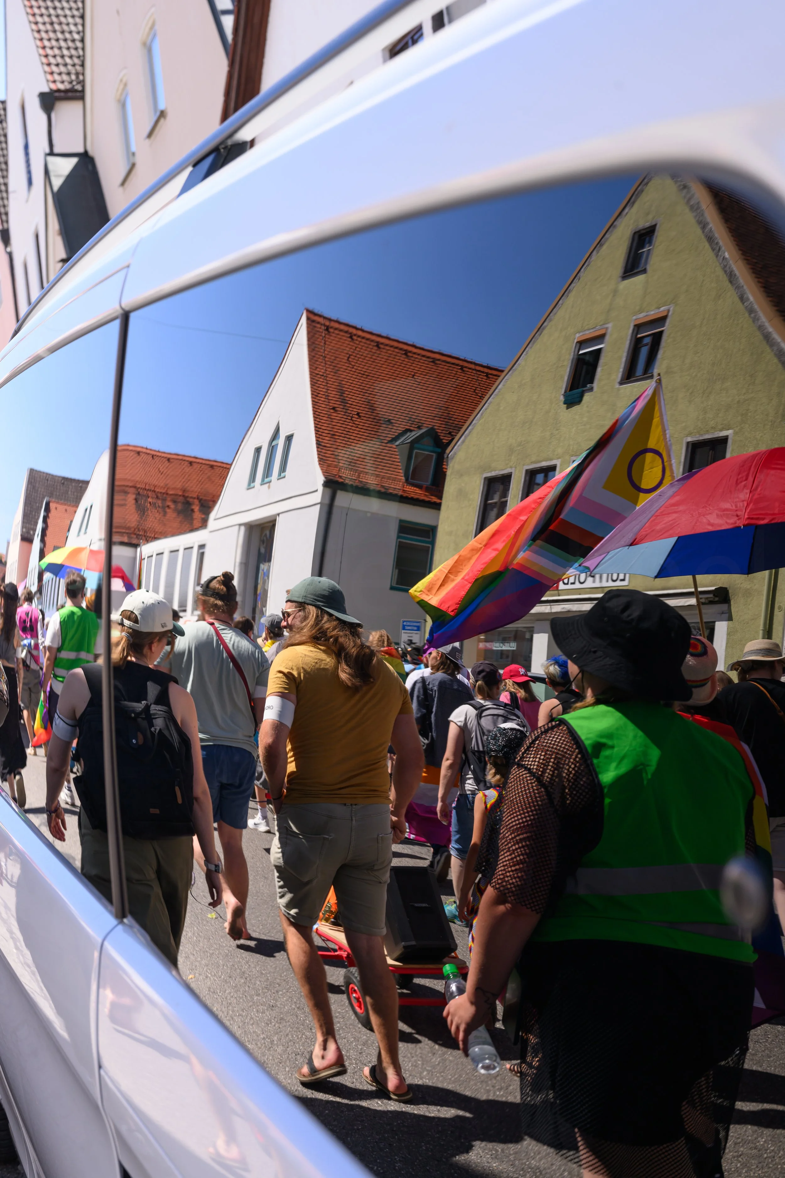 Menschen versammeln sich bei einer Parade oder Kundgebung, einige tragen Regenbogenflaggen, auf einer Straße in einer Stadt mit bunten Häusern. Es ist sonnig, viele Menschen sind in sommerlicher Kleidung.