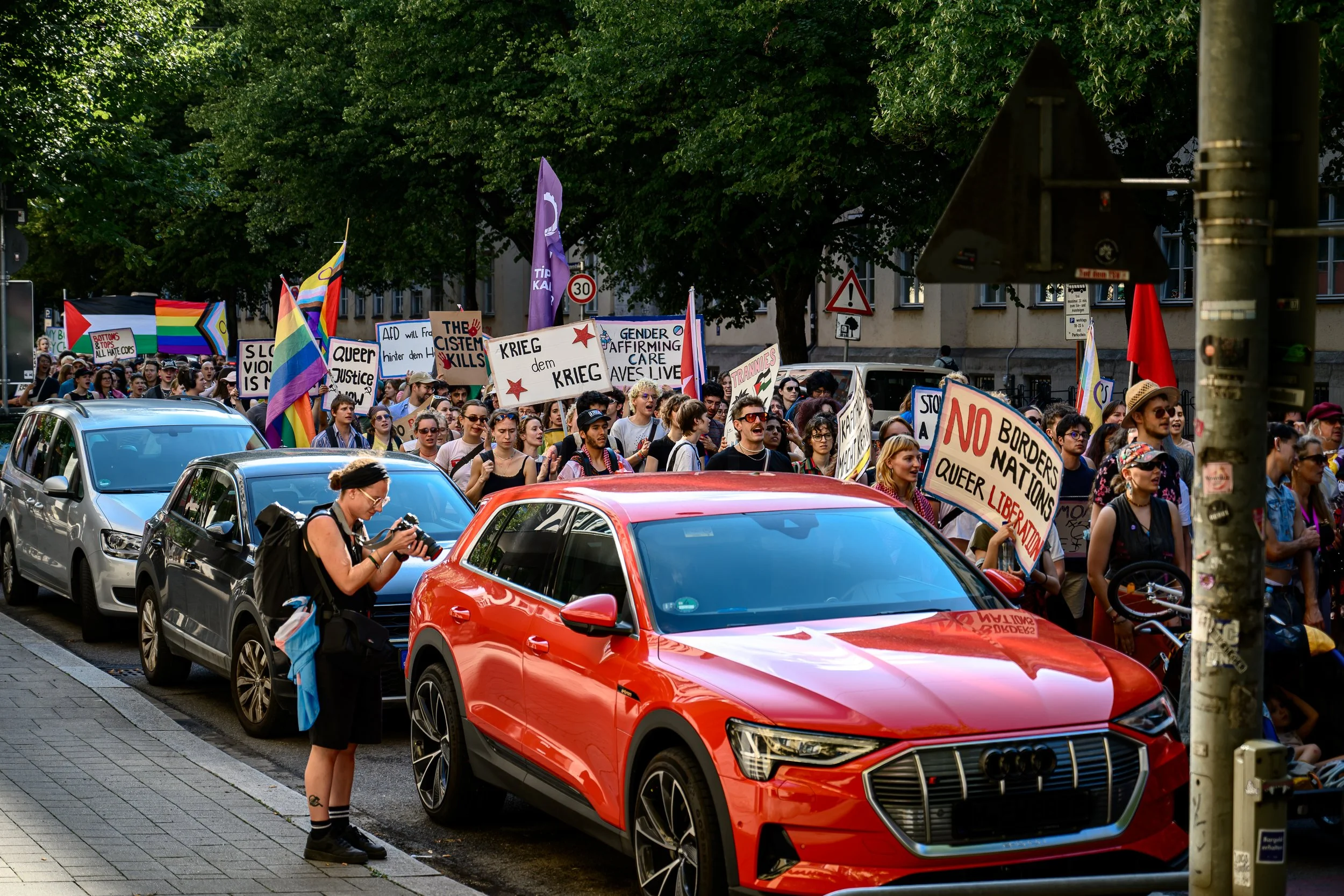 Menschen nehmen an einer Demonstration teil, die Schilder mit Frieden, Gleichstellung und LGBTQ+ Themen trägt, unterwegs auf einer Straße mit Autos und Bäumen im Hintergrund.