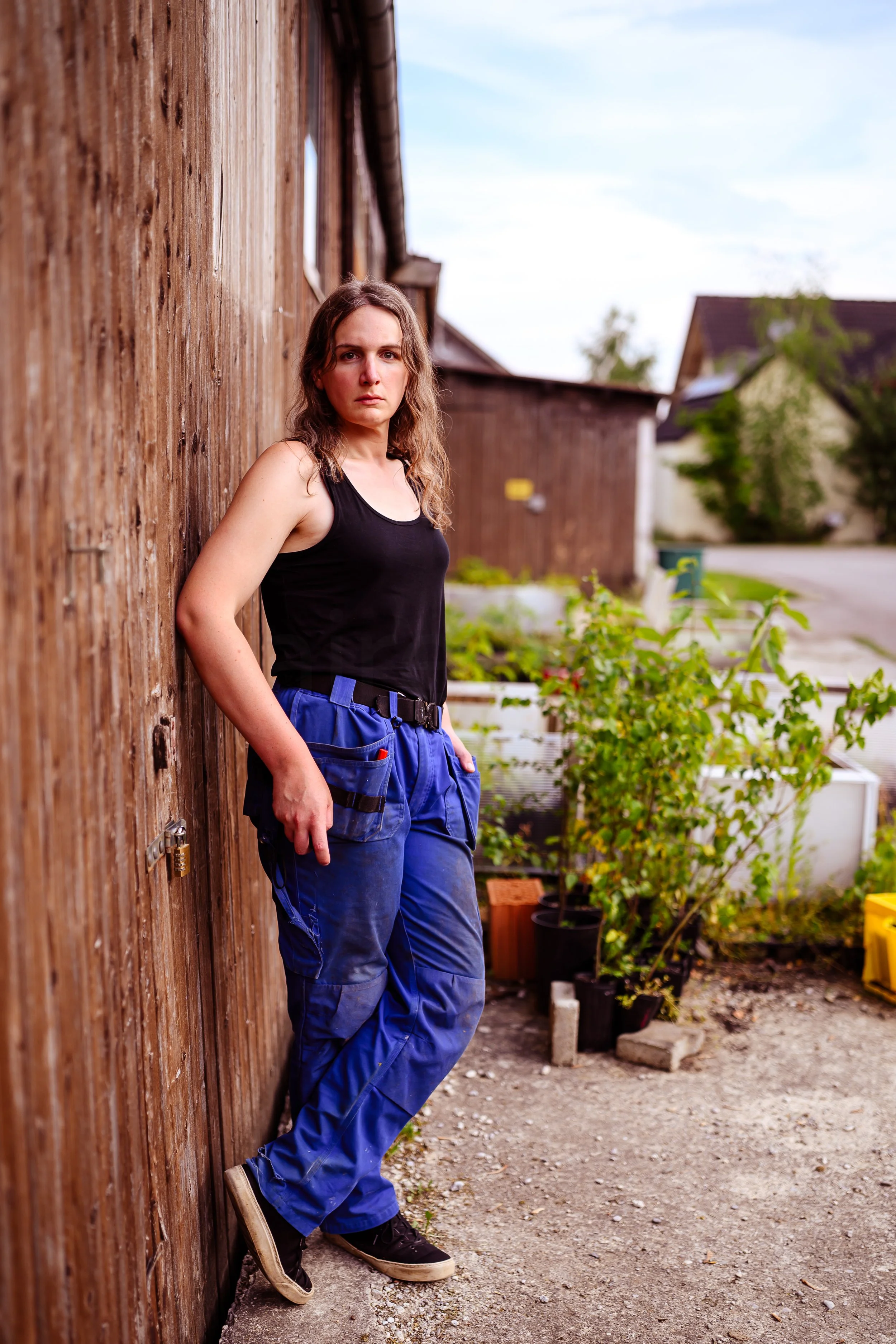 Junge Frau steht mit gesenktem Blick und verschränkten Armen an einer Holzhauswand im Freien, im Hintergrund sind mehrere Pflanzen und eine Straße sichtbar.