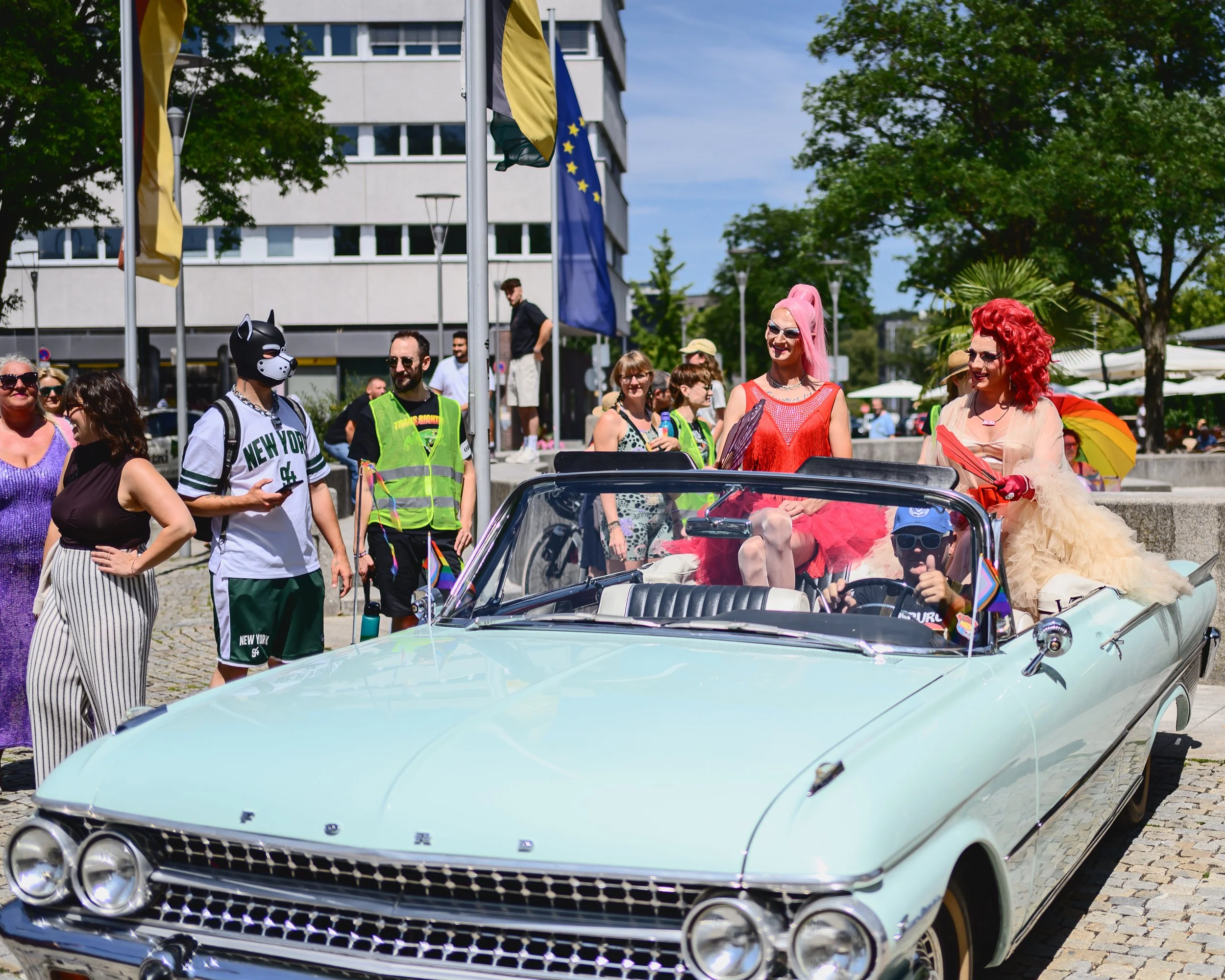Menschen bei einer Pride-Parade, einige in bunten Outfits und mit Pride-Flaggen, fahren in einem geschmückten Auto, umgeben von bunten Regenschirmen und Flaggen, im Freien mit Bäumen und modernen Gebäuden im Hintergrund.