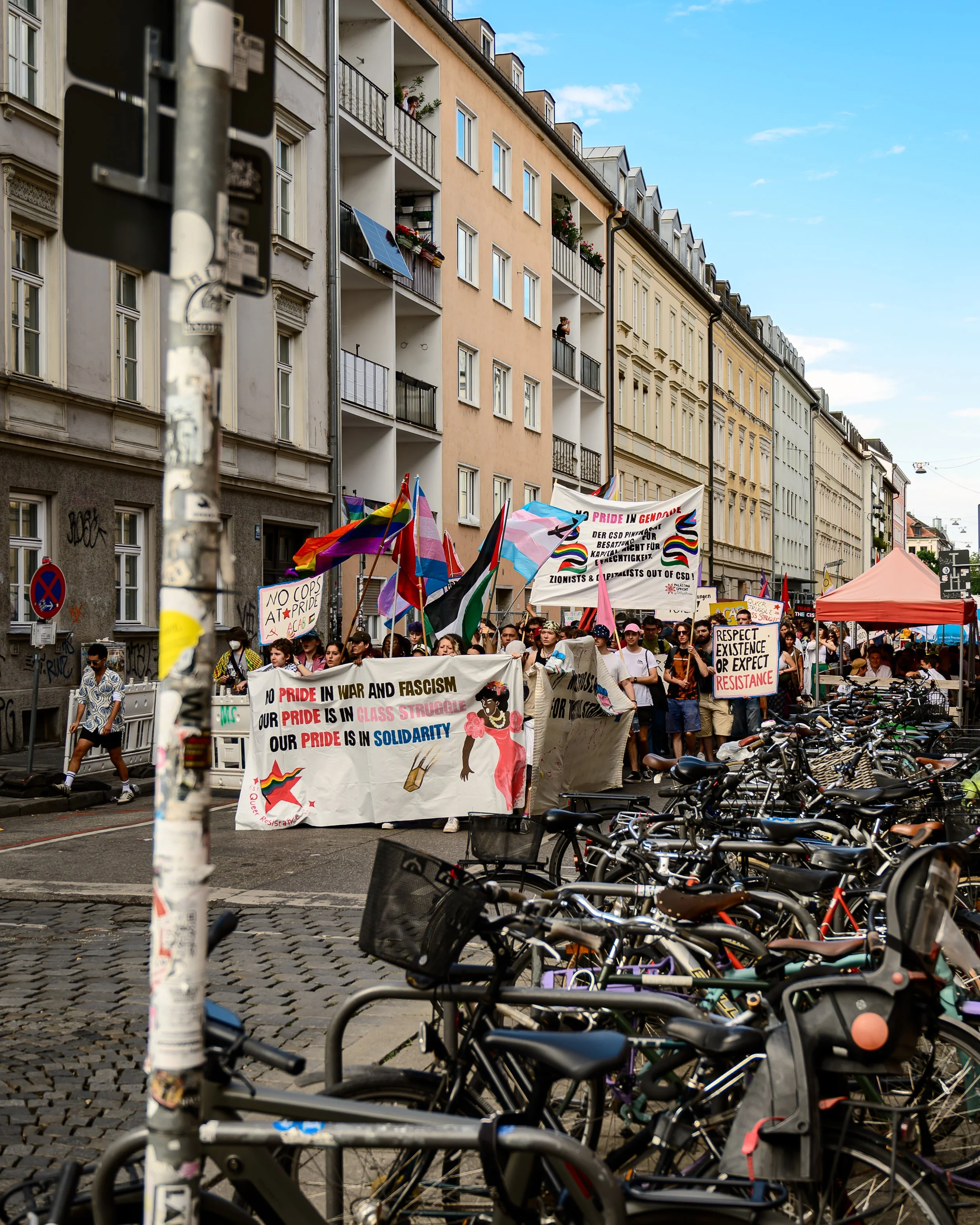 Demonstration mit Menschen, die Flaggen und Plakate tragen, in einer Stadtstraße mit mehrstöckigen Häusern im Hintergrund, Fahrradparkplätze im Vordergrund.