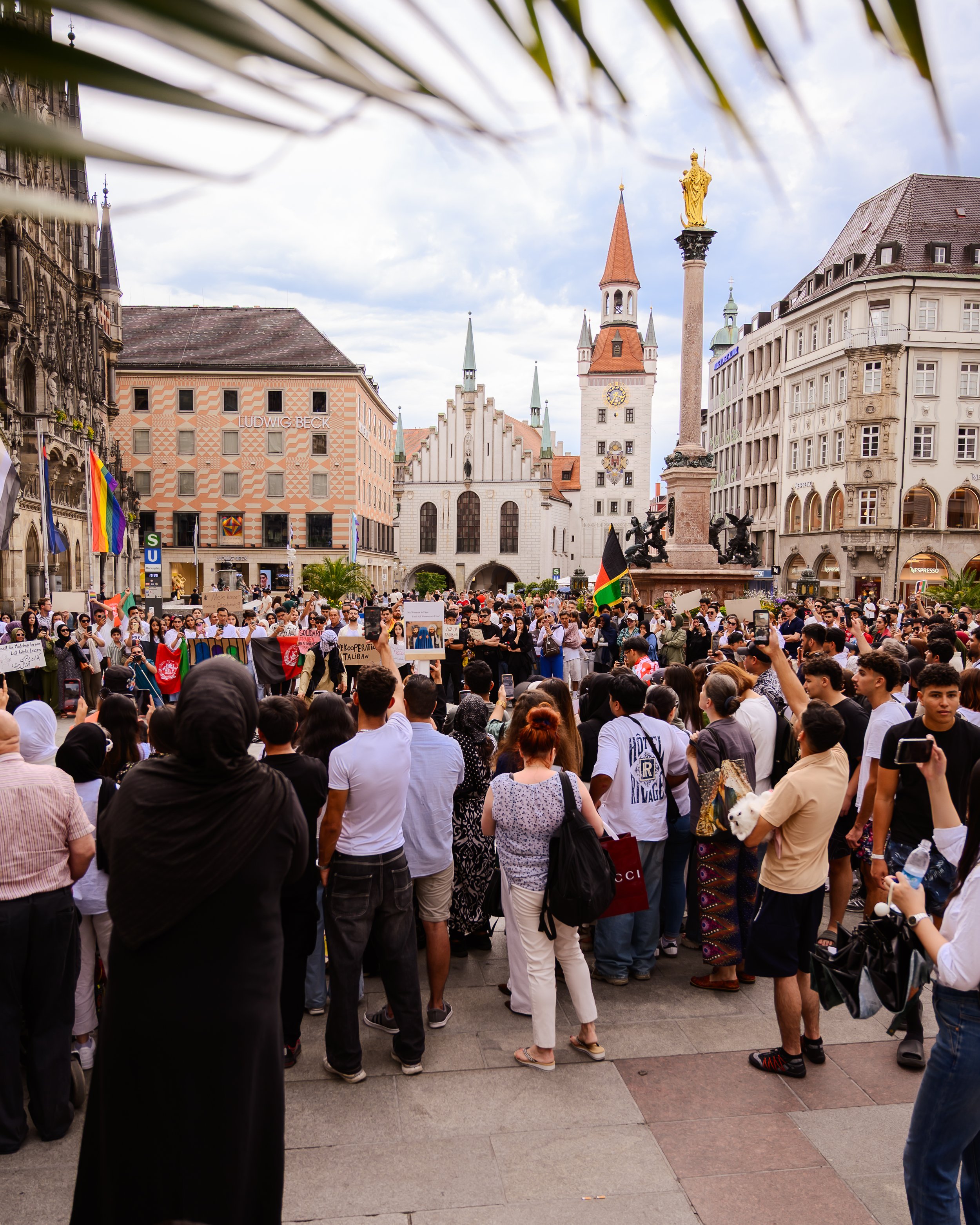 Eine große Menschenmenge versammelt sich auf einem Platz in einer Stadt, mit historischen Gebäuden im Hintergrund, darunter eine Kirche und Denkmäler, während einige Menschen Schilder und Fahnen halten.