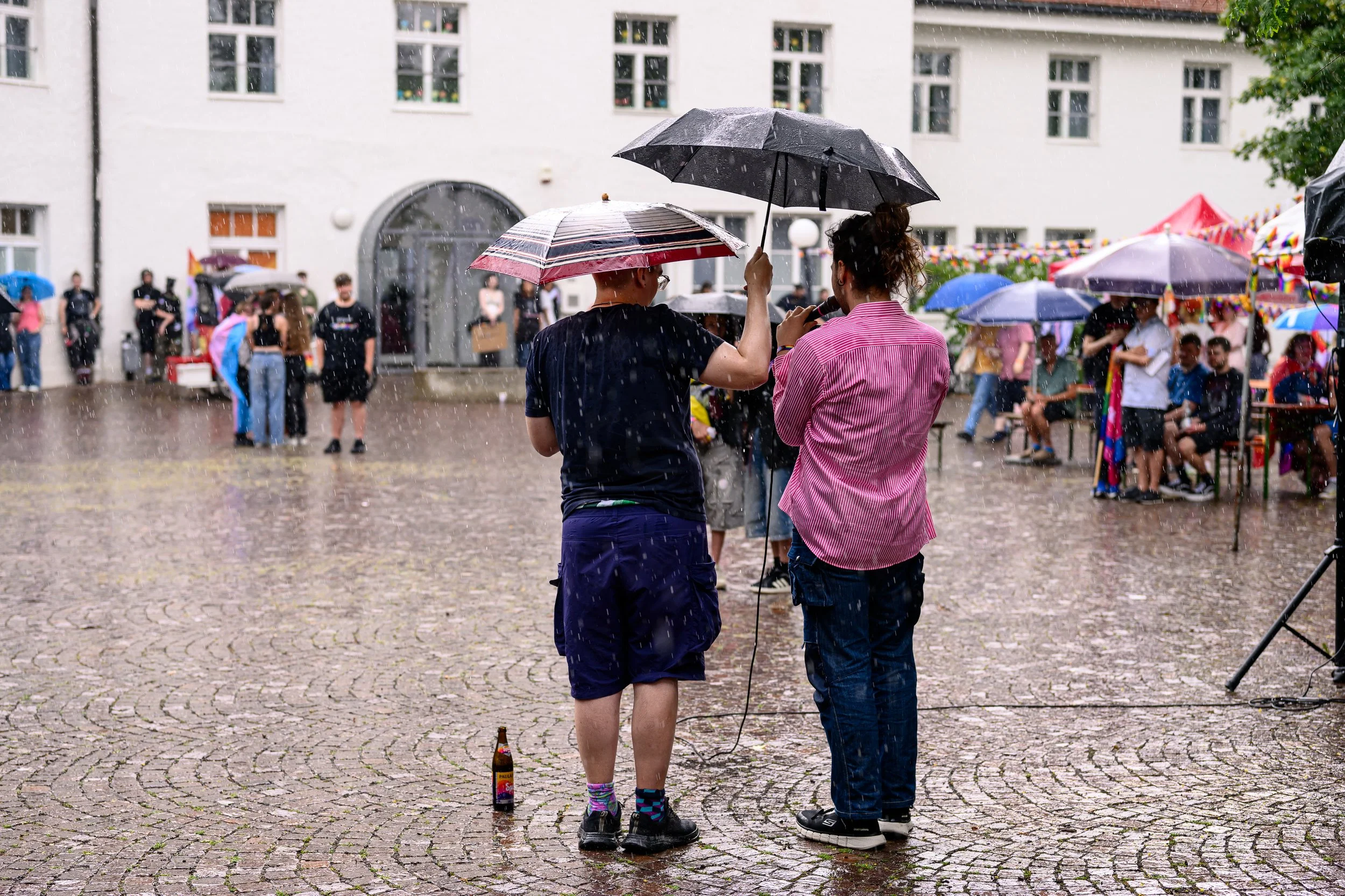 Zwei Menschen mit Regenschirmen unter Regen auf einem gepflasterten Platz, im Hintergrund eine Gruppe von wartenden Menschen und ein Gebäude.