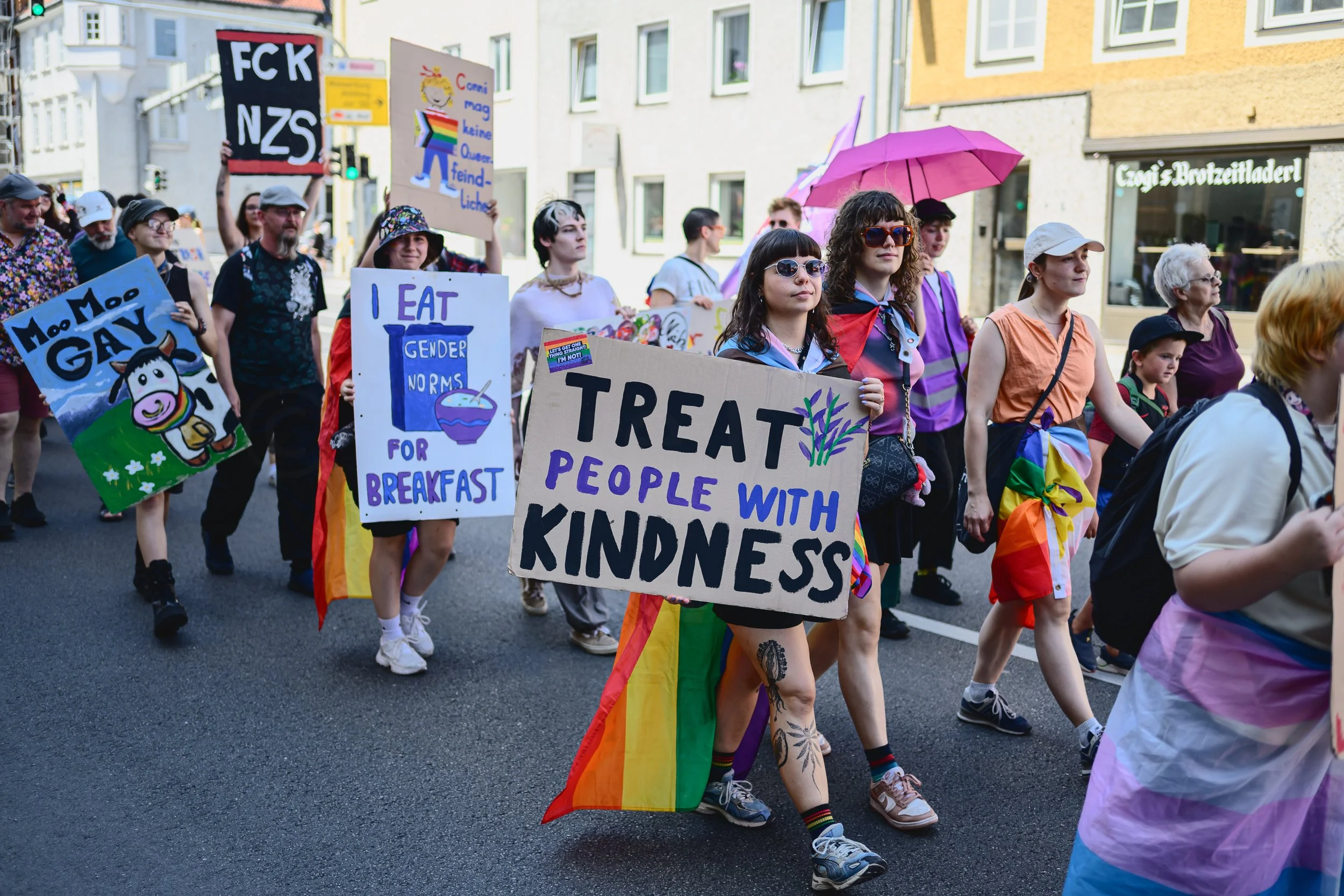 Eine Gruppe von Menschen läuft bei einer Demonstration oder Parade. Sie halten Schilder mit Botschaften für Gleichberechtigung, Vielfalt und Akzeptanz, einige tragen bunte Kleidung und Regenmäntel. Im Vordergrund ein Schild mit der Aufschrift 'Treat 