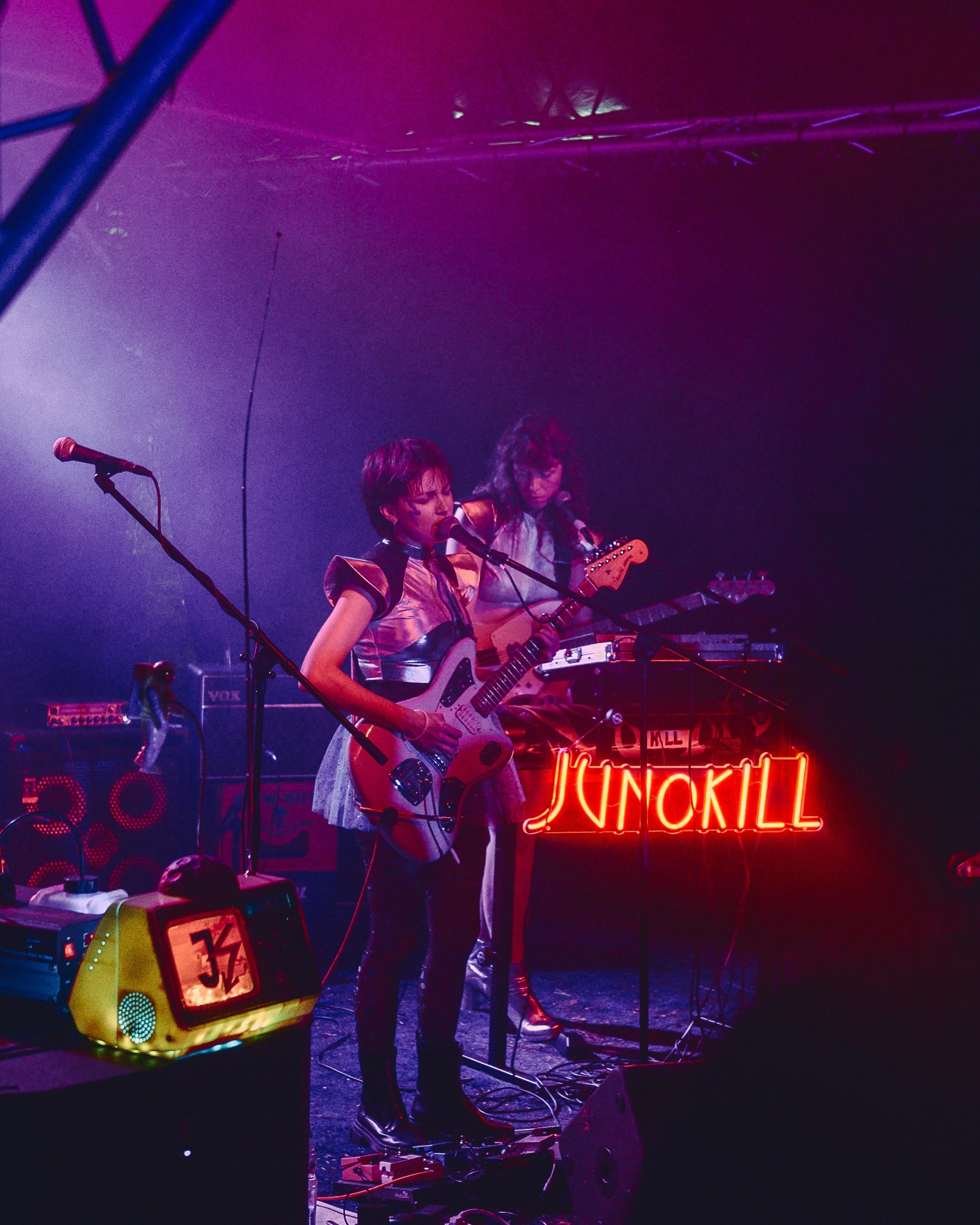 Zwei Musikerinnen mit Gitarren spielen auf einer Bühne, umgeben von Lichteffekten und Musiktechnik, mit einer Neonbeschriftung im Hintergrund.