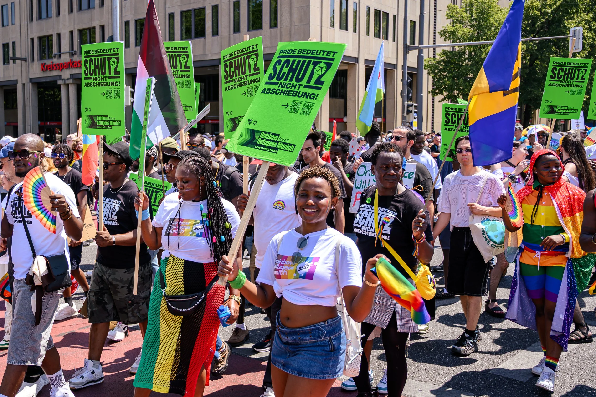 Menschen bei einer Regenbogenparade demonstrieren mit Regenbogenfahnen, bunten Kleidern und Schildern, die für LGBTQ+ Rechte und gegen Diskriminierung sind.