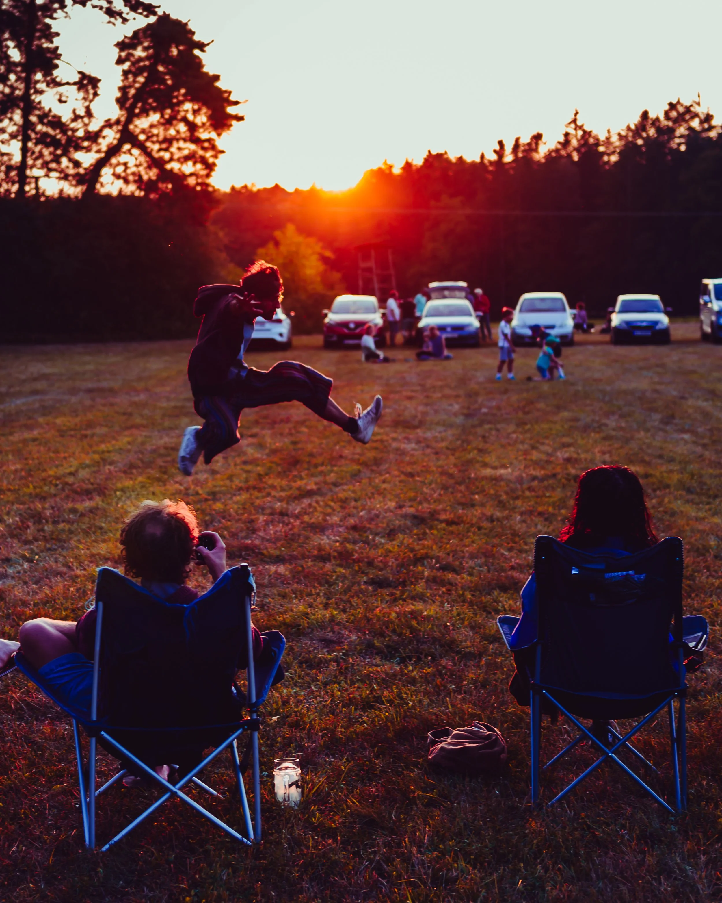 Sonnenuntergang über einer Gruppe von Menschen, die im Freien sitzen und Kinder, die spielen. Einige sitzen in Campingstühlen, andere stehen oder sind bei Aktivitäten. Es gibt Fahrzeuge im Hintergrund. Die Szene ist ruhig und abseits der Stadt.