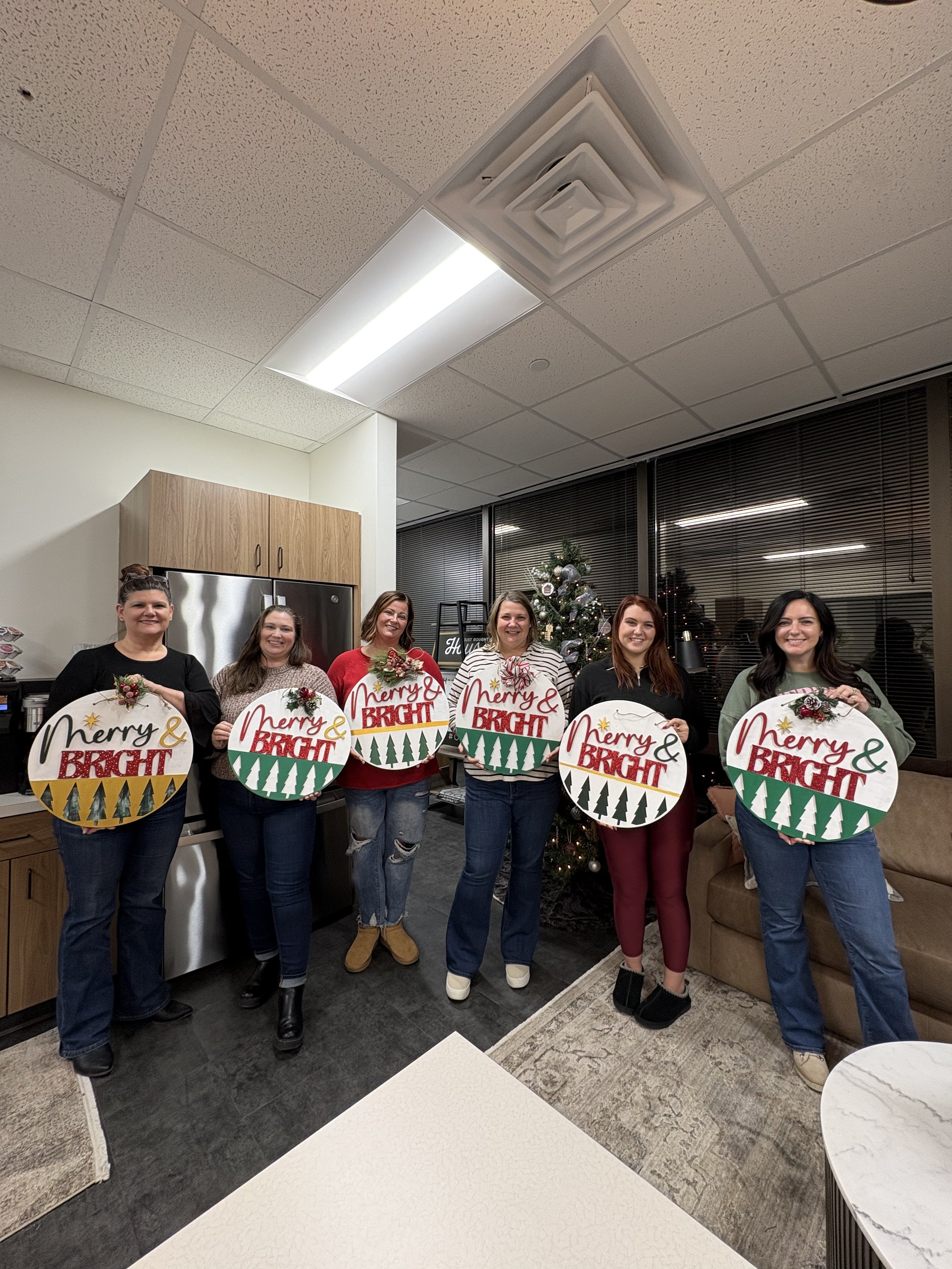 Two women smiling and talking at a painting event, with art supplies and canvases on display in the background. They are wearing blue aprons.