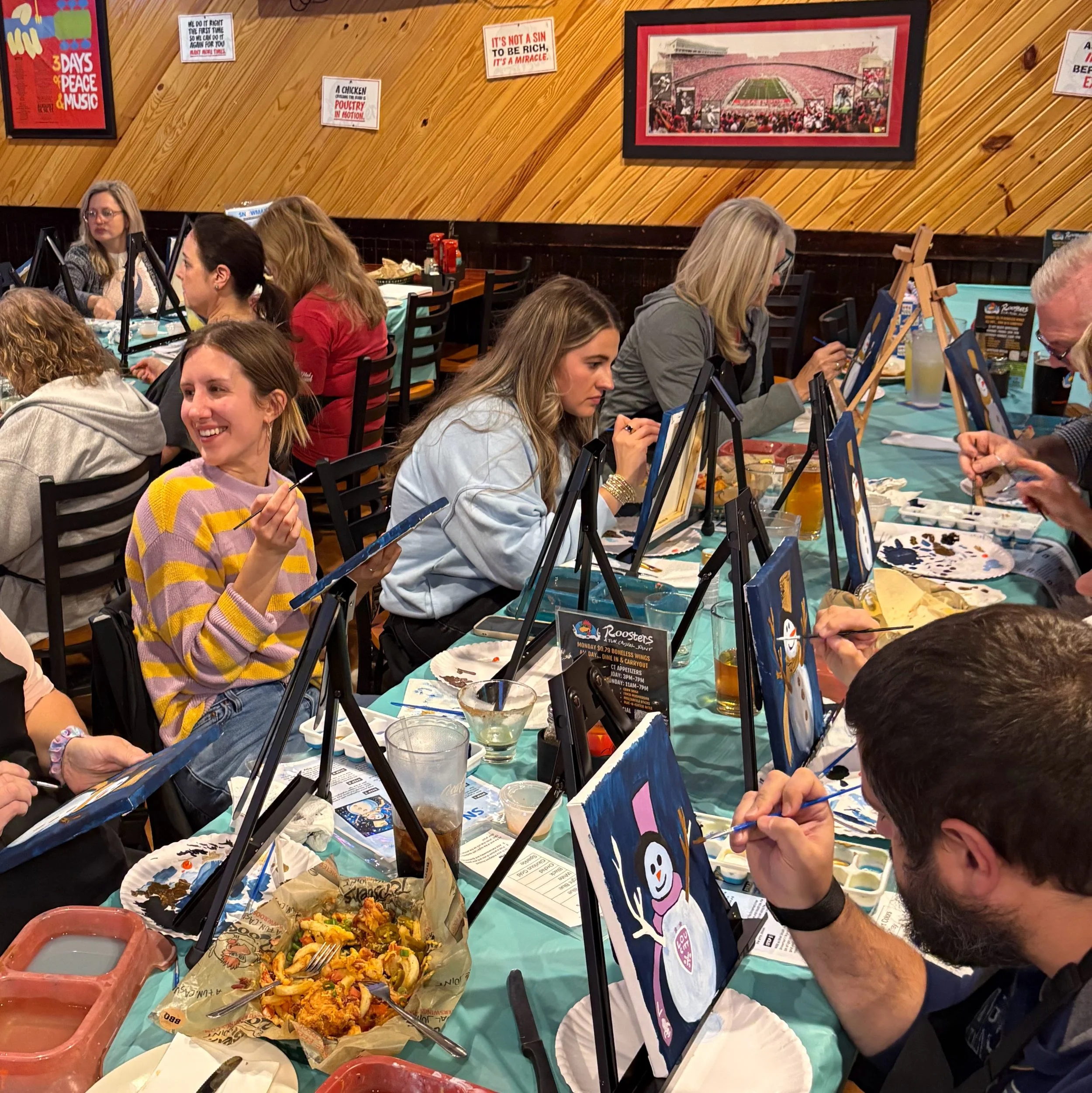 Two women smiling and talking at a painting event, with art supplies and canvases on display in the background. They are wearing blue aprons.