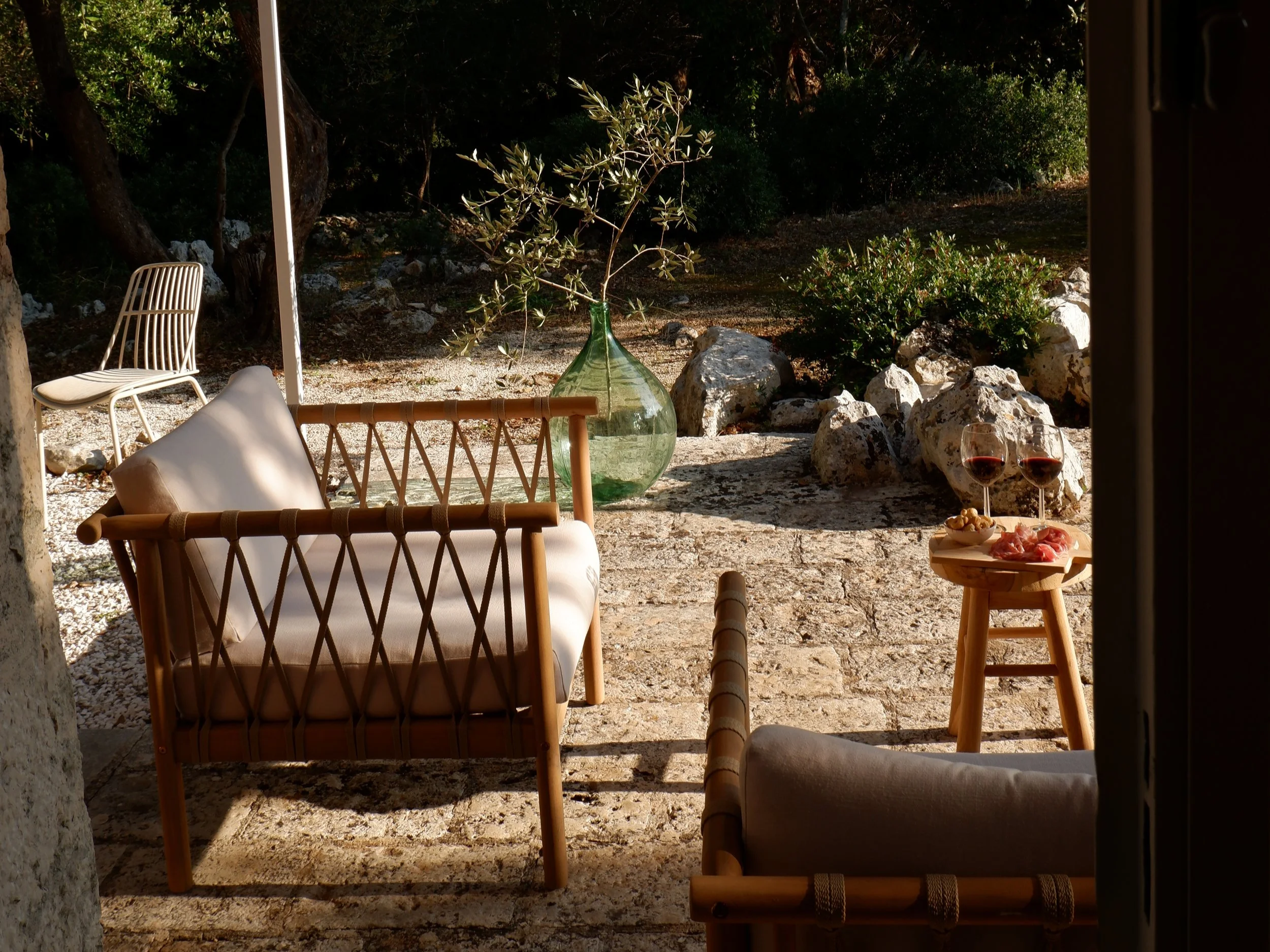 Uitzicht op een patio met houten en stoffen meubels, een grote groene vaas met een tak, een tafeltje met glazen wijn en charcuterie, omgeven door stenen en groen