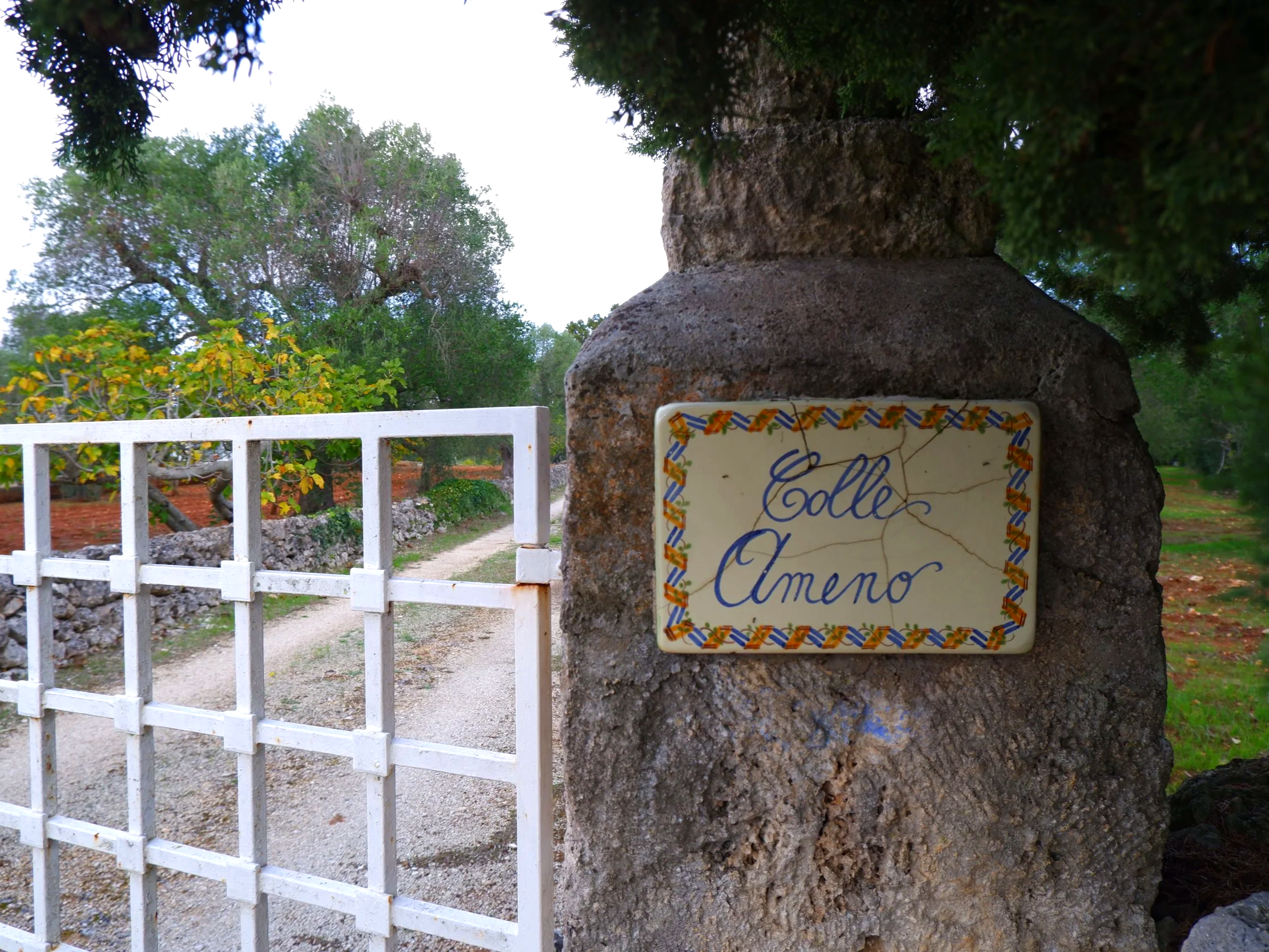 Uitsnede van een stenen poort met een keramische naamplaat met de tekst 'Colle Amene' op een landweg, omgeven door groen en bomen.
