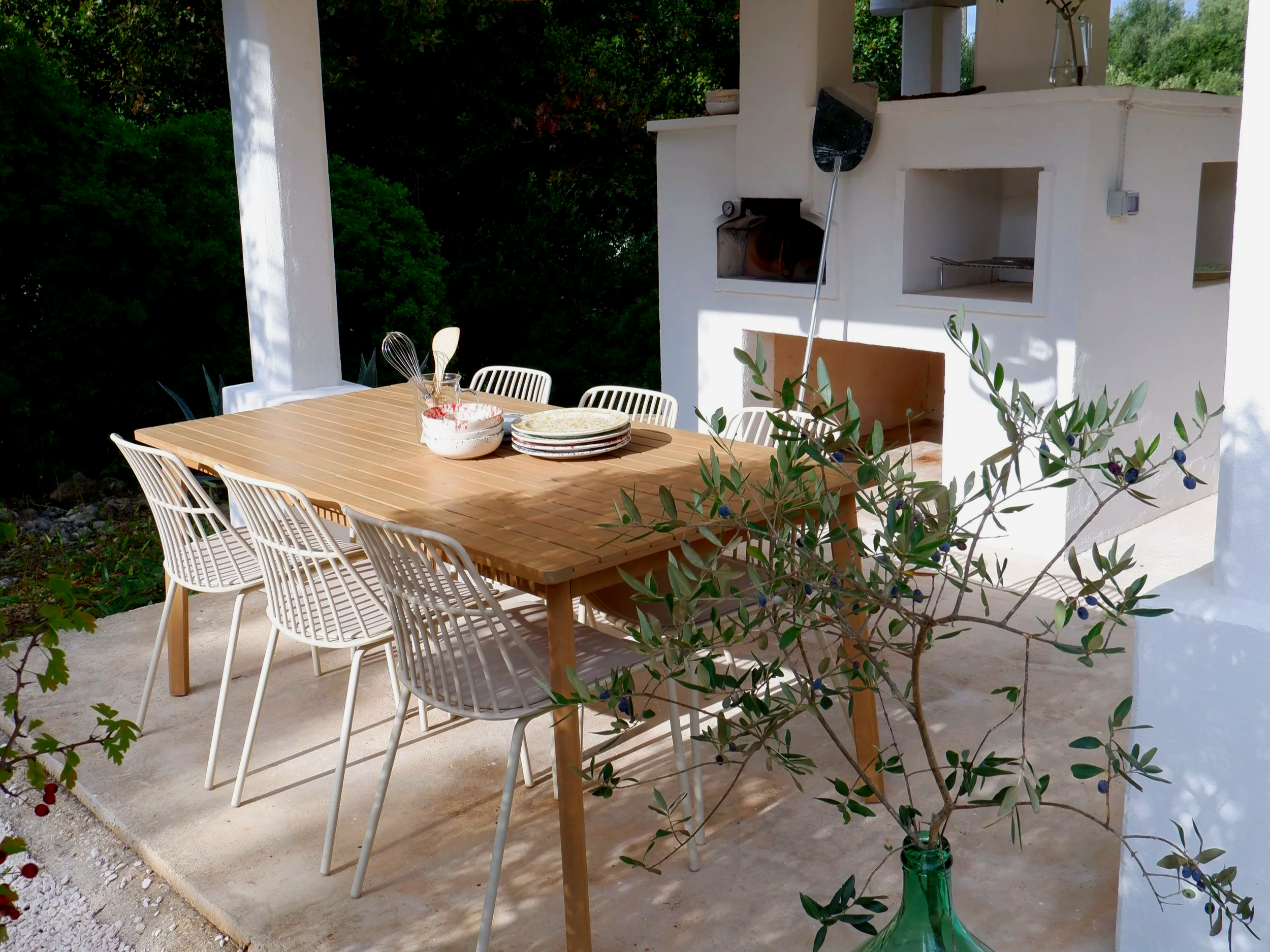 Een buitentafel met acht witte stoelen, een boerderijvuurplaats, planten en een vaatwasser in de muur, en een groen bladvormige olijfboom met bessen in een groene fles.