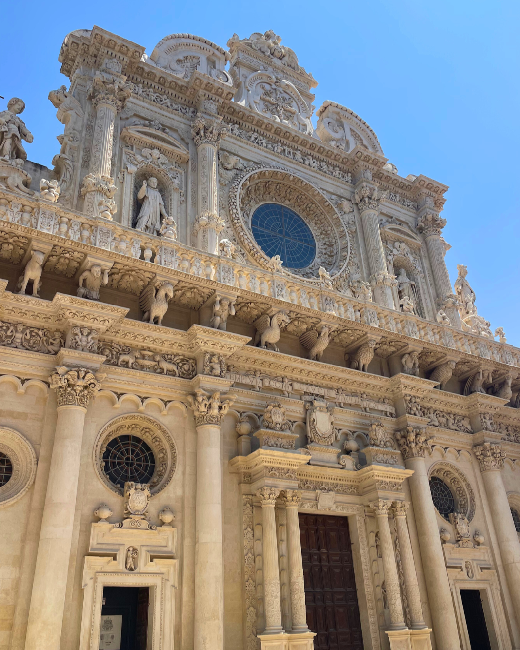 Een gedetailleerde barokke kerkgevel gemaakt van wit marmer met beeldhouwwerken, kolommen en decoratieve elementen onder een heldere blauwe hemel.