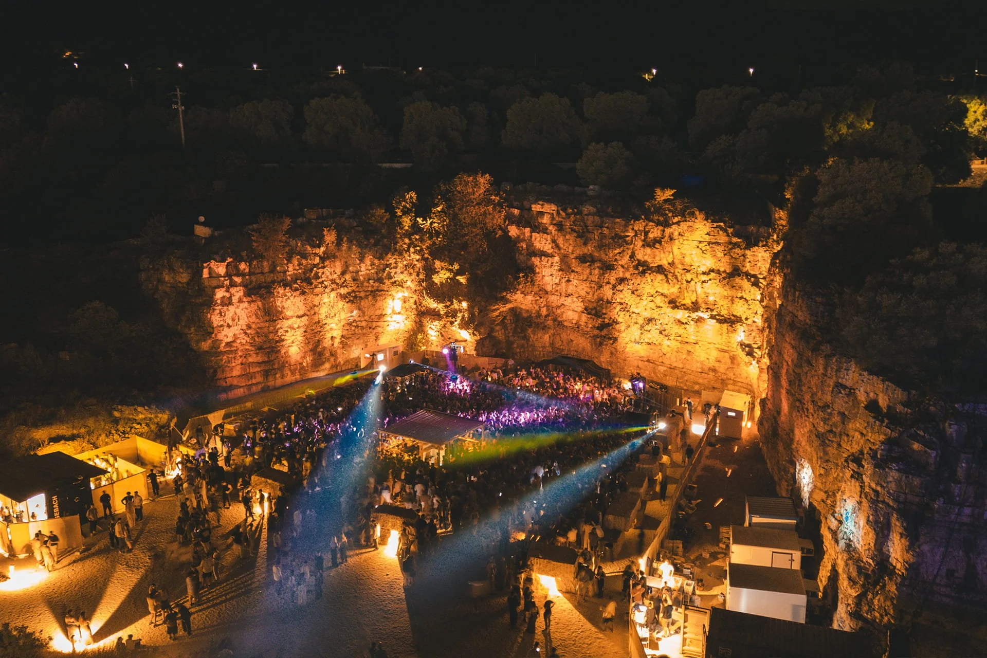 Feest op een locatie met rotswanden en een dansvloer, verlicht door kleurrijke lichten en vol mensen.