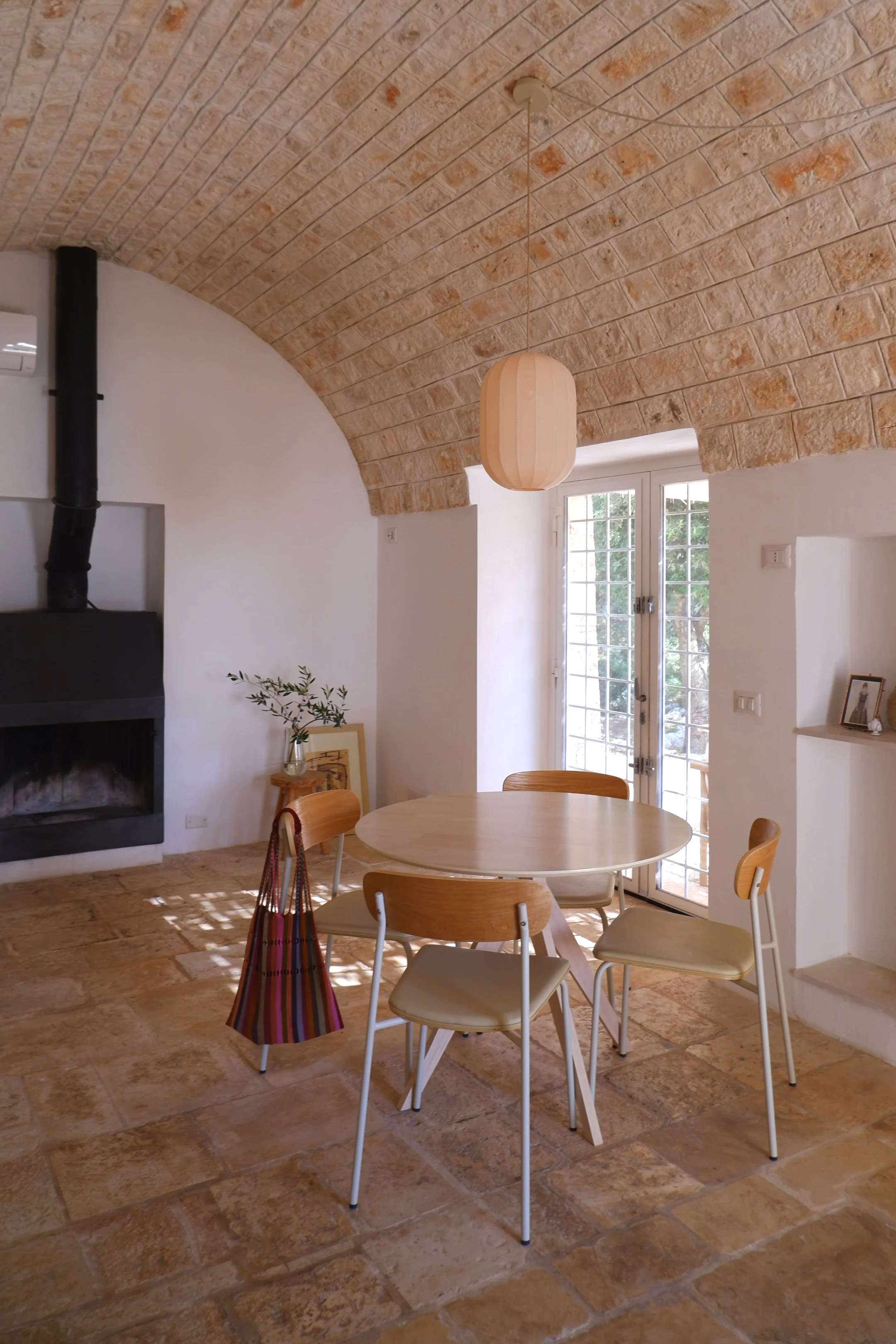 Interieur van een kamer met een stenen gebogen plafond, een ronde houten eettafel met vier houten stoelen, een open haard, een glazen deur naar buiten, en decoraties zoals een plant en een foto.