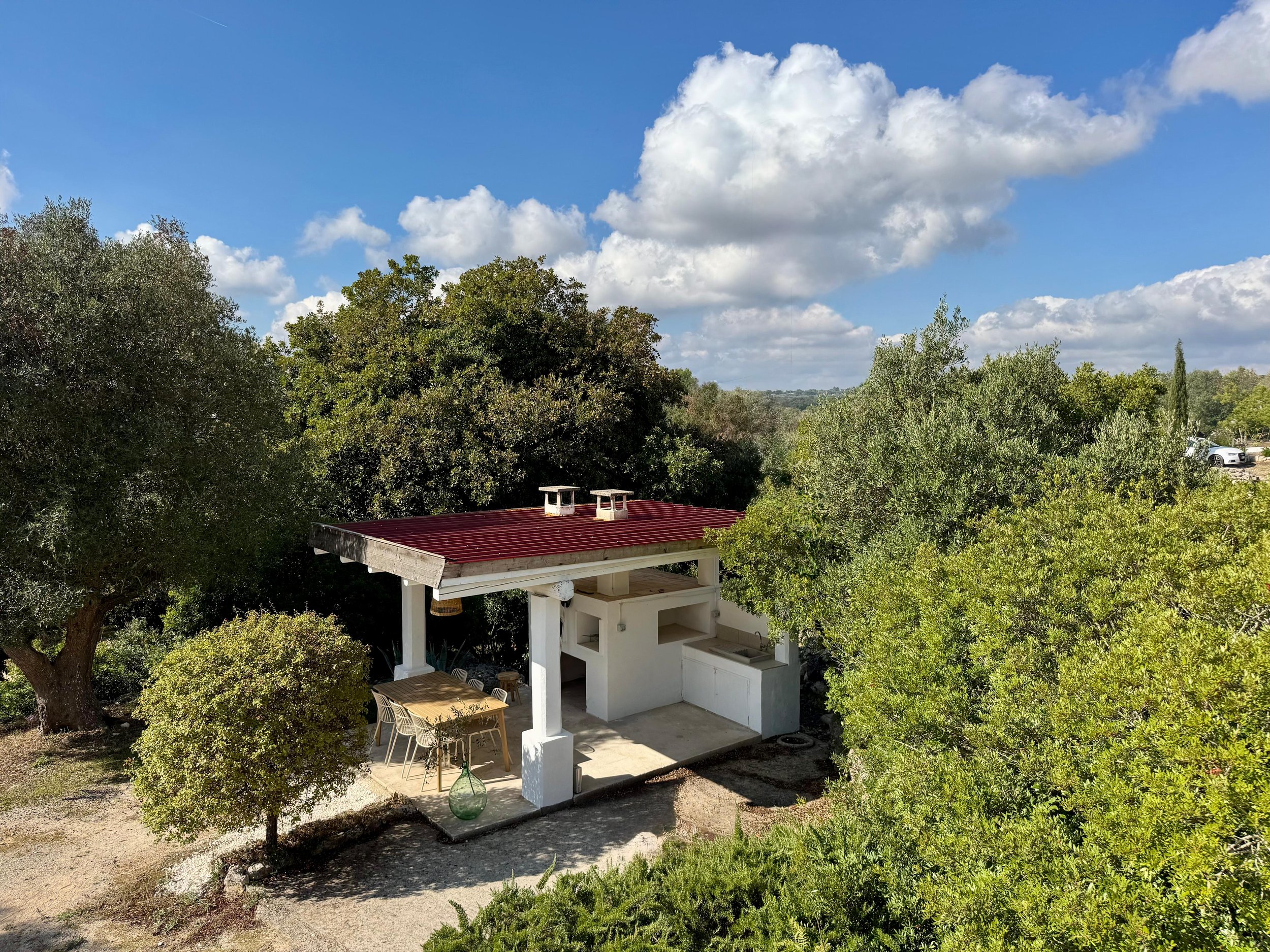 Buitenkeuken met witgebouwd fornuis, rode dak en ronde tafel met stoelen, omringd door groene bomen onder een bewolkte blauwe hemel.