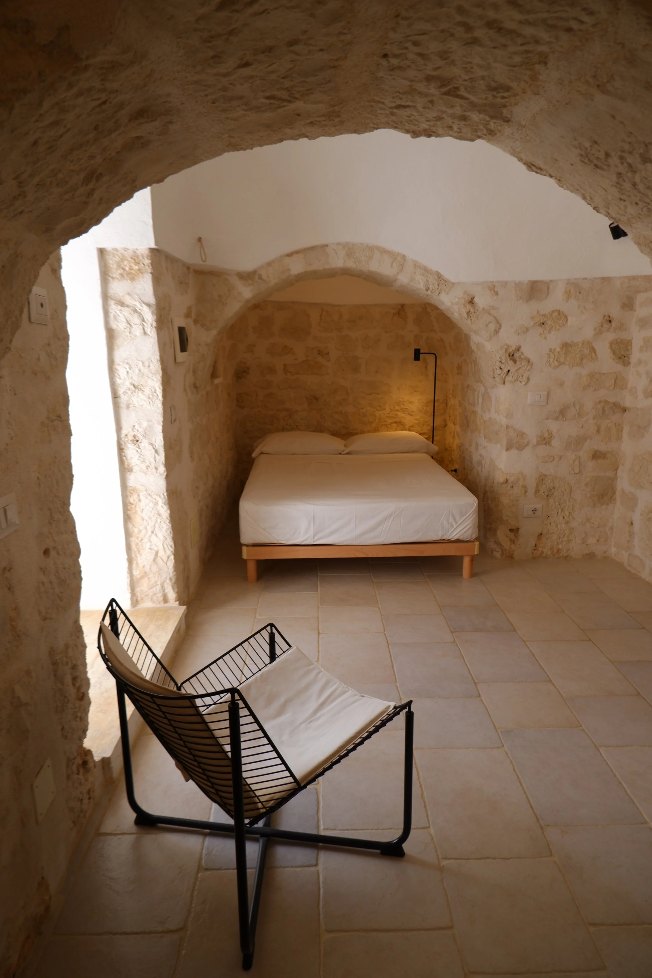 Een slaapkamer met stenen muren en plafonds, een bed met witte lakens, en een zwart metalen stoel in de voorgrond.