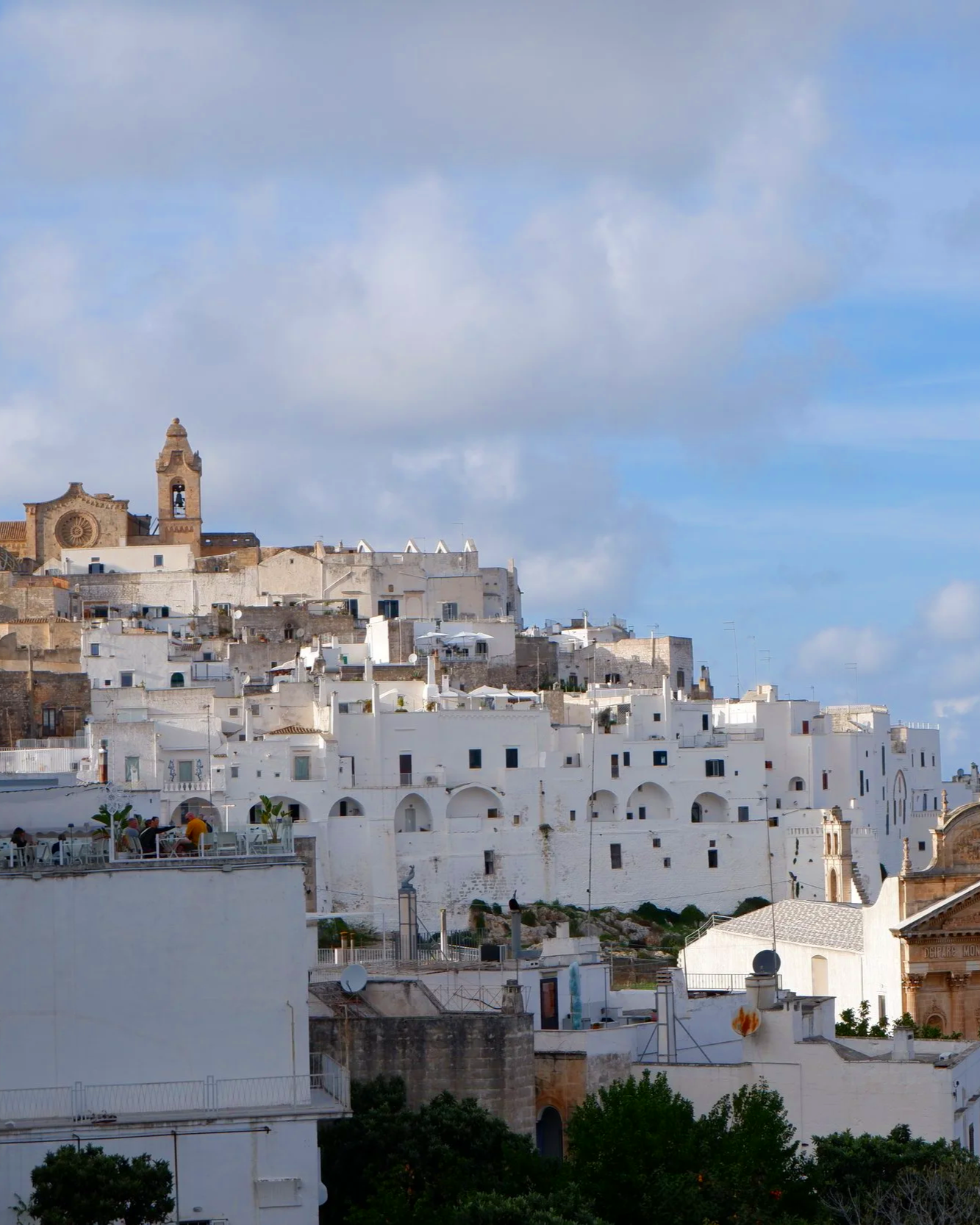 Uitzicht op een witte stad met veel witte gebouwen op een heuvel, met een kerk met een toren en een blauwe lucht met wolken op de achtergrond.