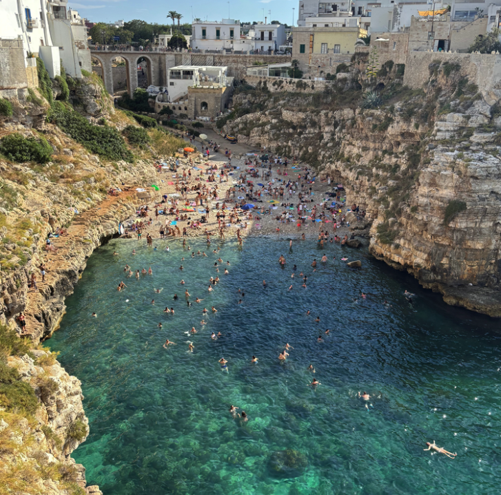 Een druk strand met veel mensen die zwemmen en zonnen, omringd door rotsen en een stad met witte gebouwen op de achtergrond.