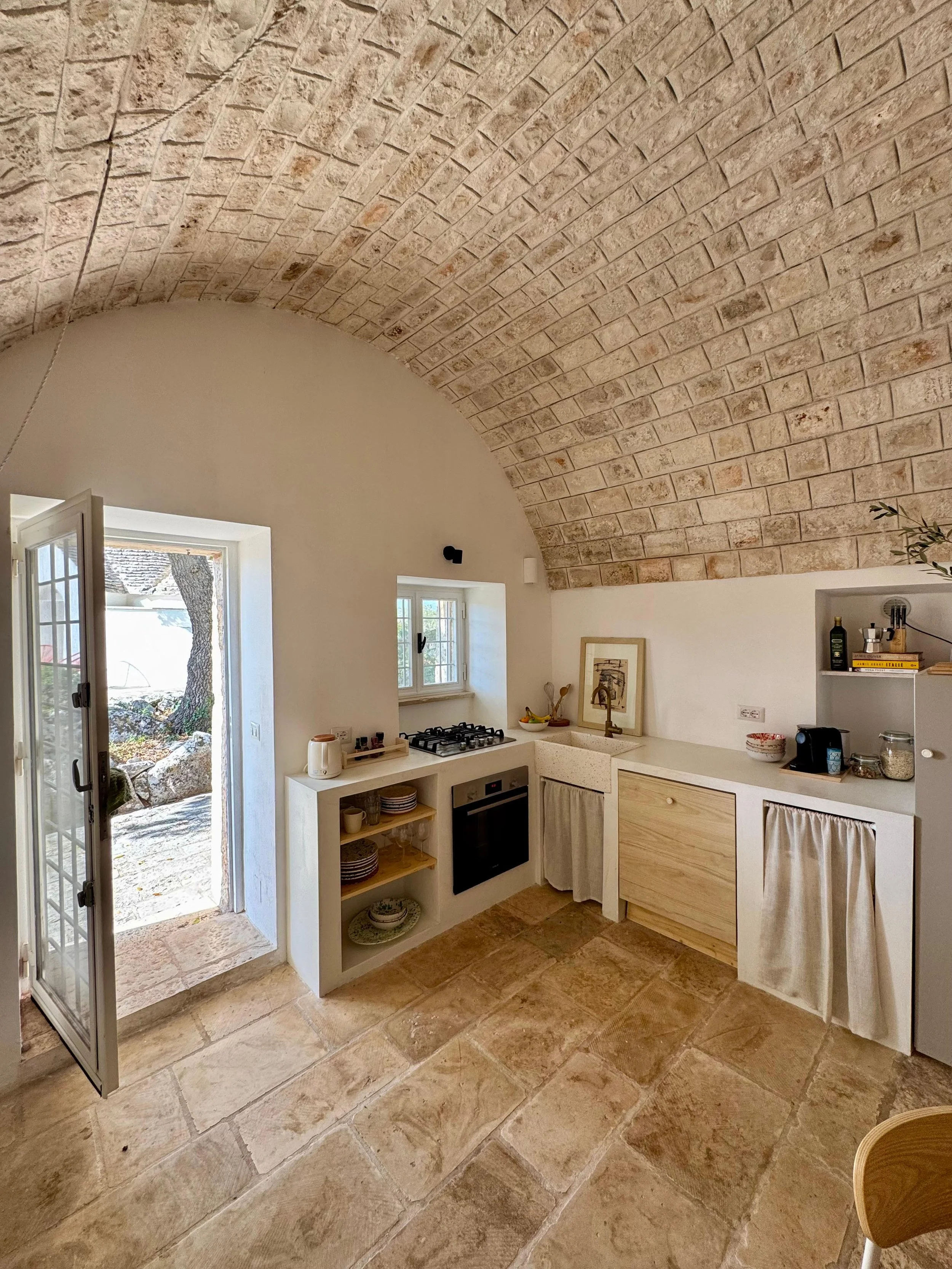 Een keuken met een gebogen bakstenen plafond, een open deur naar buiten, en witte kasten met gordijntjes onder de werkbladen. Er is een vierkant raam, een fornuis, en verschillende keukengerei en decoraties.