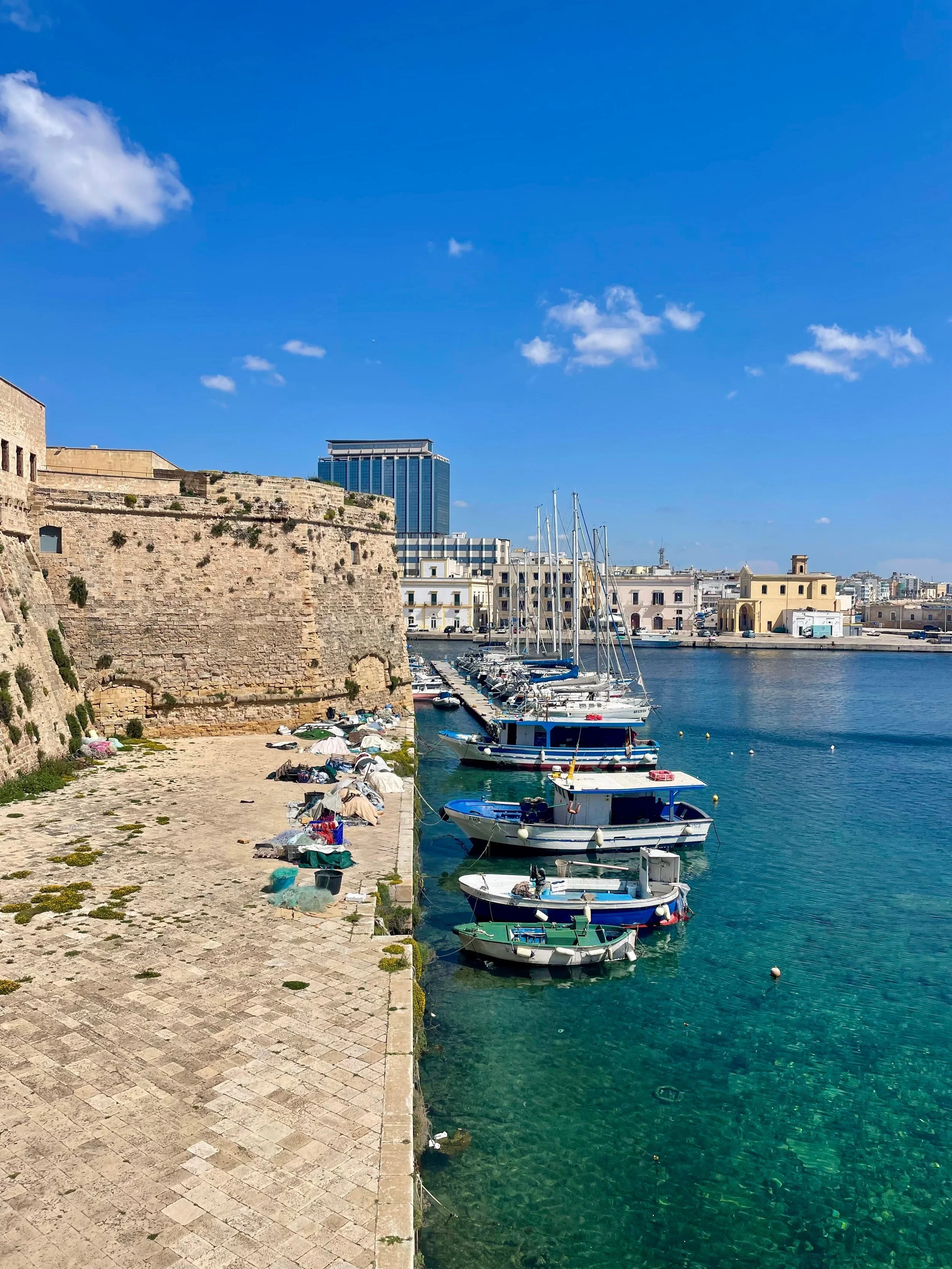 Een zonnige haven met ligplaatsen voor boten, een oude muur aan de linkerkant, moderne gebouwen op de achtergrond, en een blauwe hemel met enkele witte wolken.