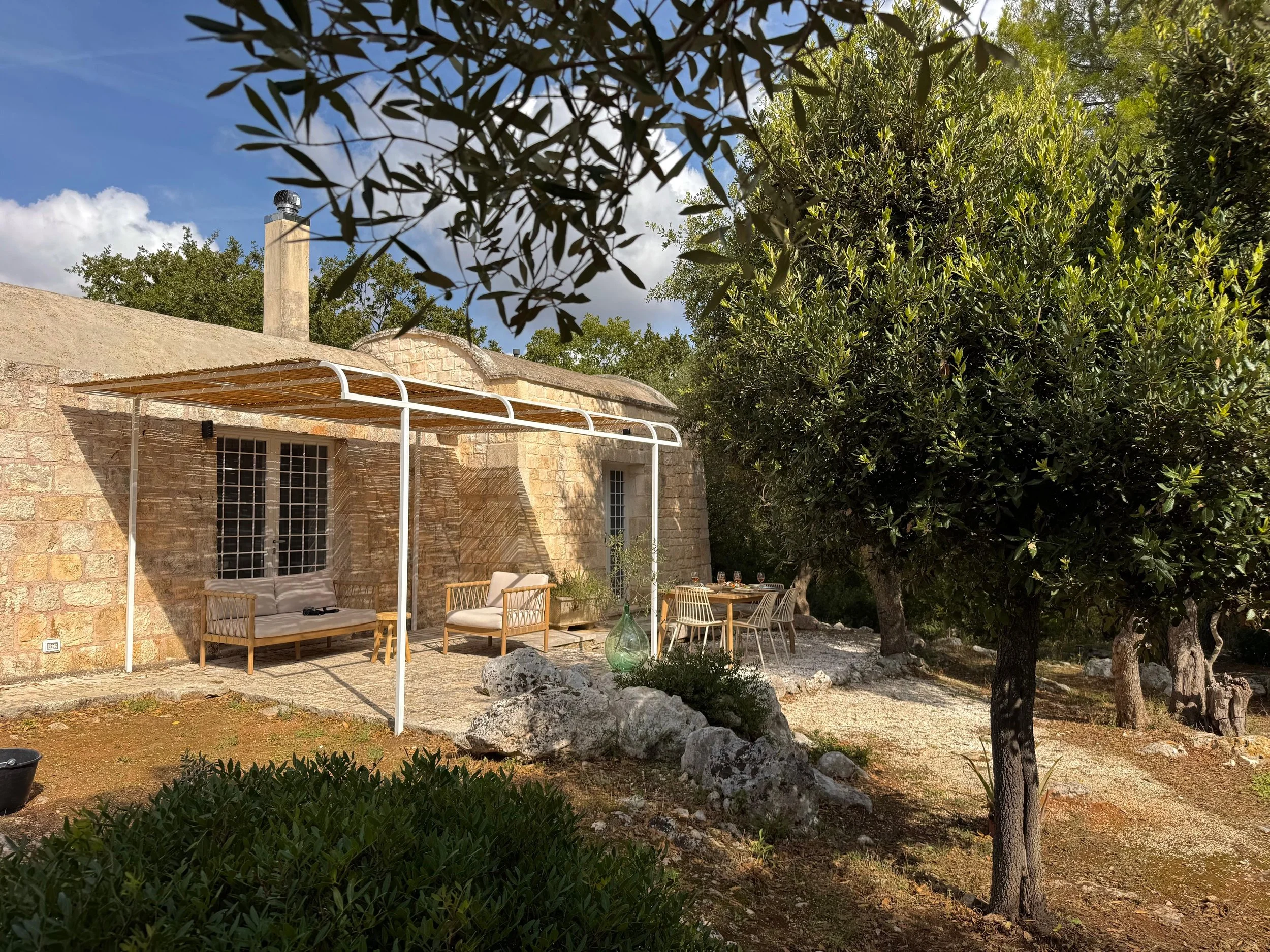 Een landelijke tuin met een stenen huis, georganiseerde zitgelegenheid onder een witte pergola, en een tuin met enkele bomen en planten.