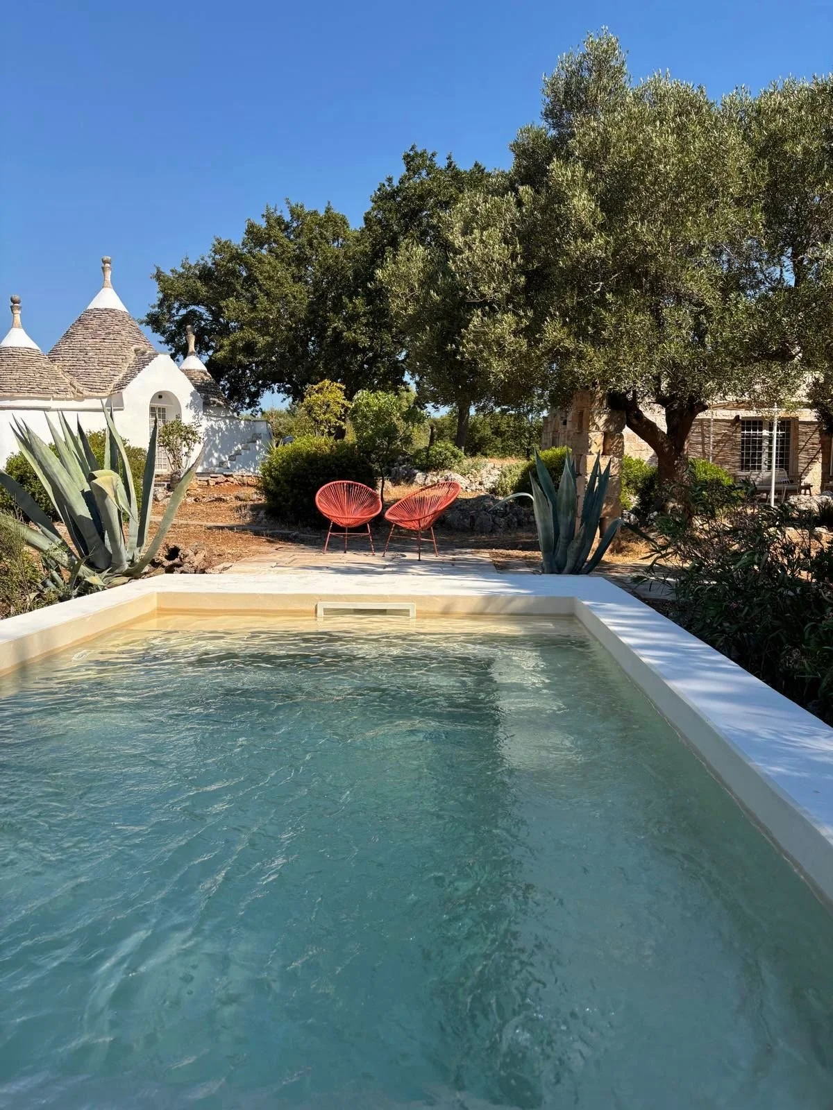 Zwembad op een terras met twee rode ligstoelen, omringd door planten en bomen, met witte gebouwen in de achtergrond, onder een heldere blauwe hemel.