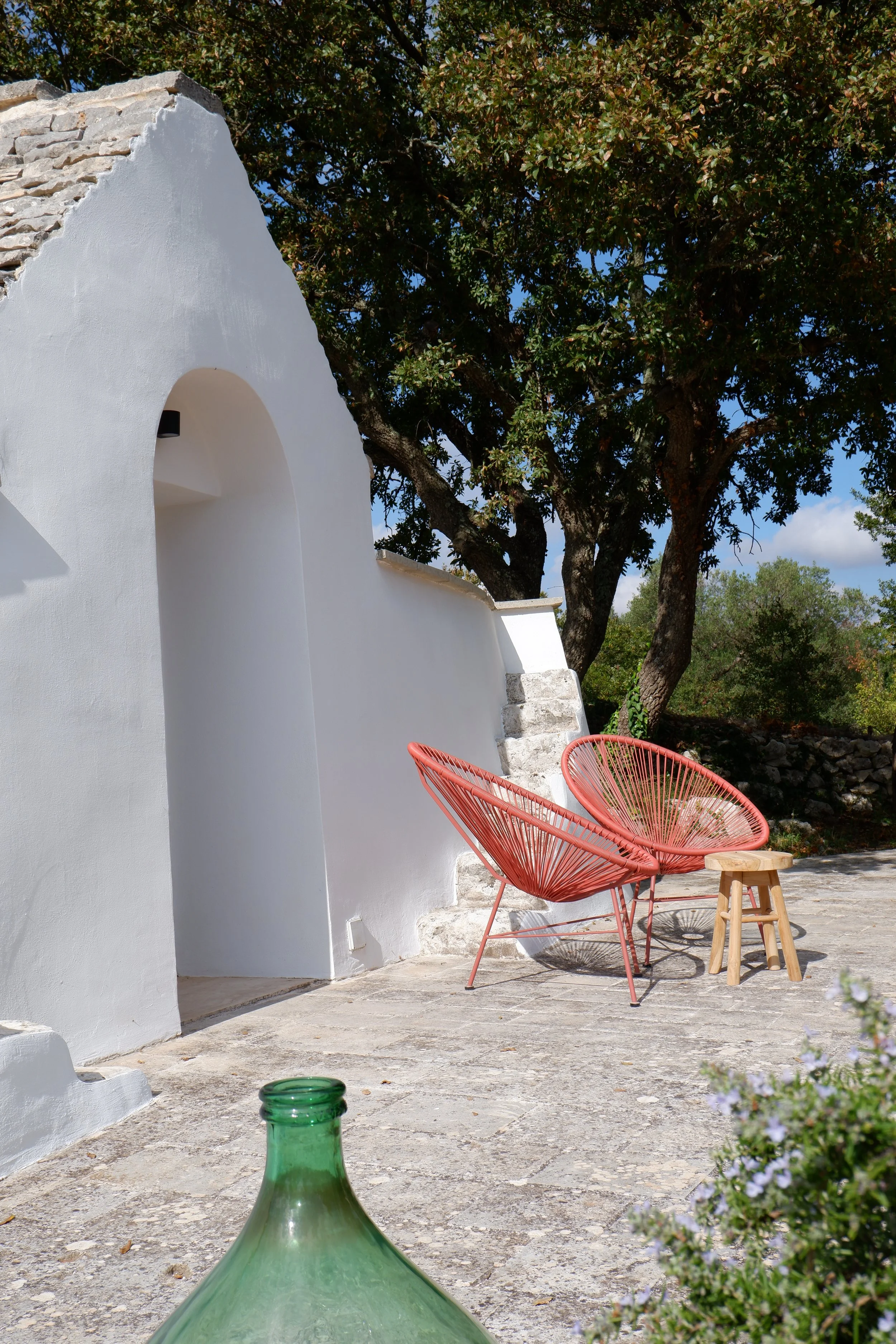 Een witte muur met een boog en tuinmeubilair bestaande uit twee rode acapulco-stoelen en een klein houten krukje, rondom een boom in een zonnige tuin.