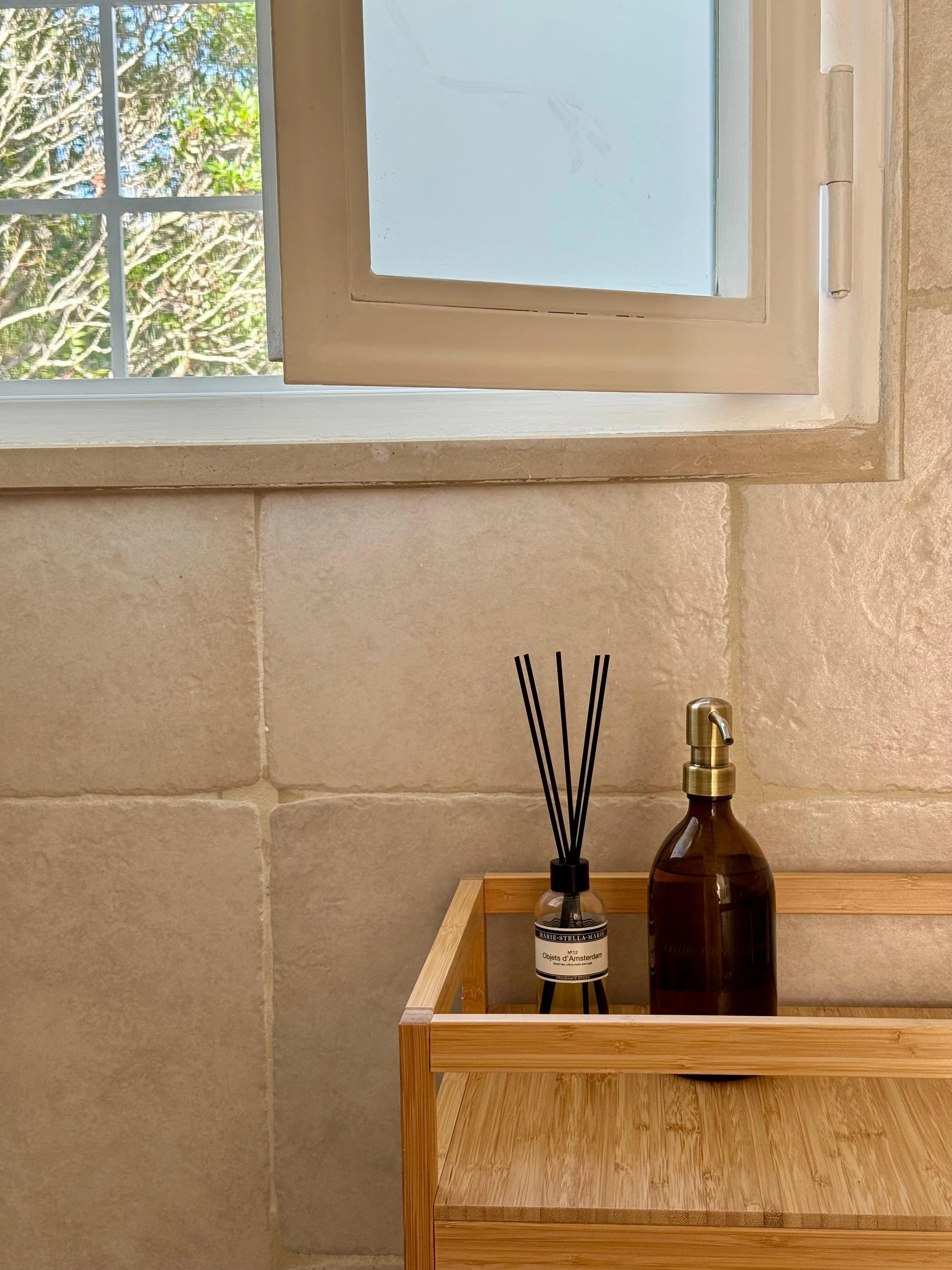 Een venster met een open raam en een houten kastje met een luchtverfrisser en een dispenser op een beige stenen muur.