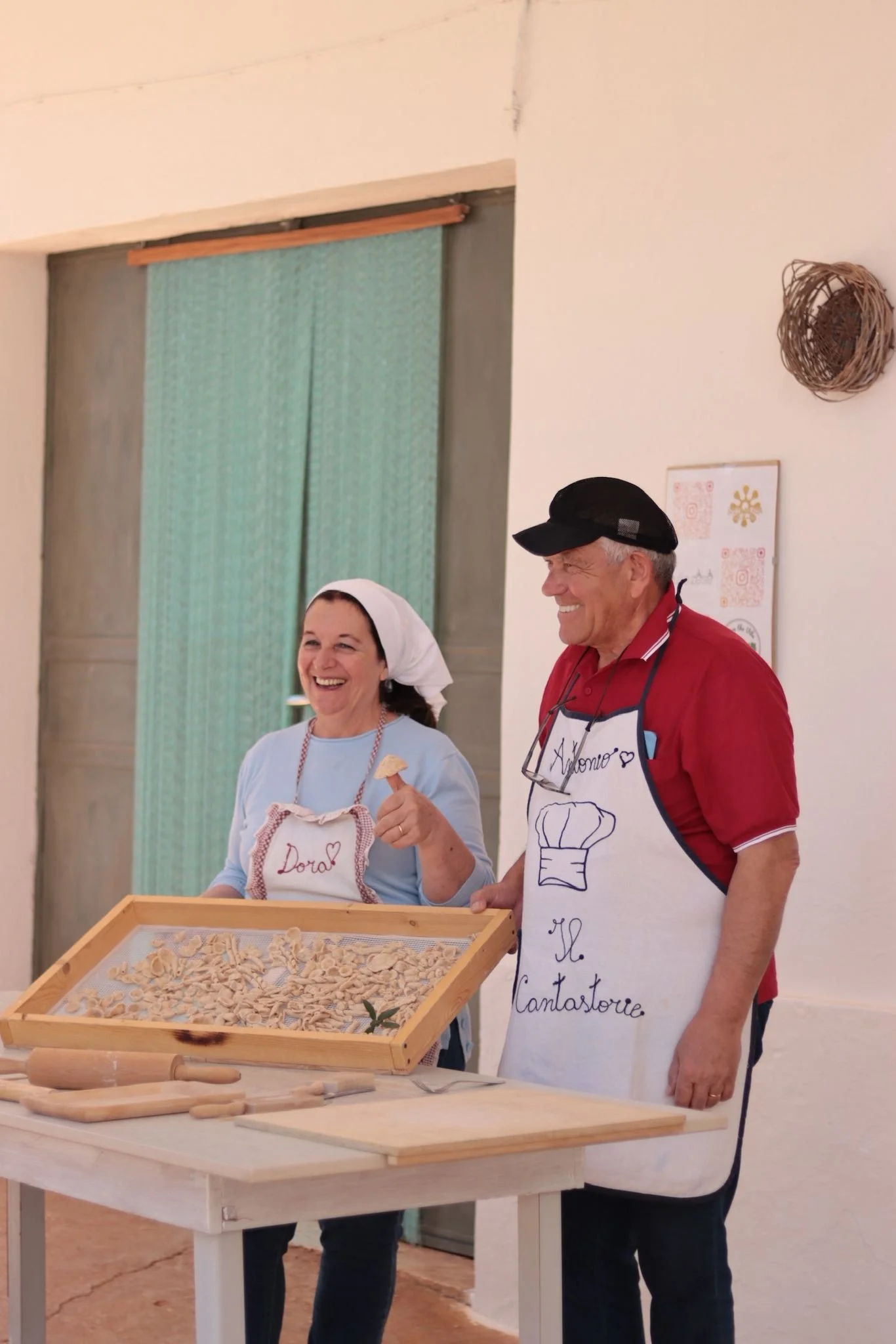 Twee mensen, een vrouw en een man, lachen terwijl ze samen in een keuken of werkruimte actief zijn met pasta maken. Ze dragen aprons en lachen.