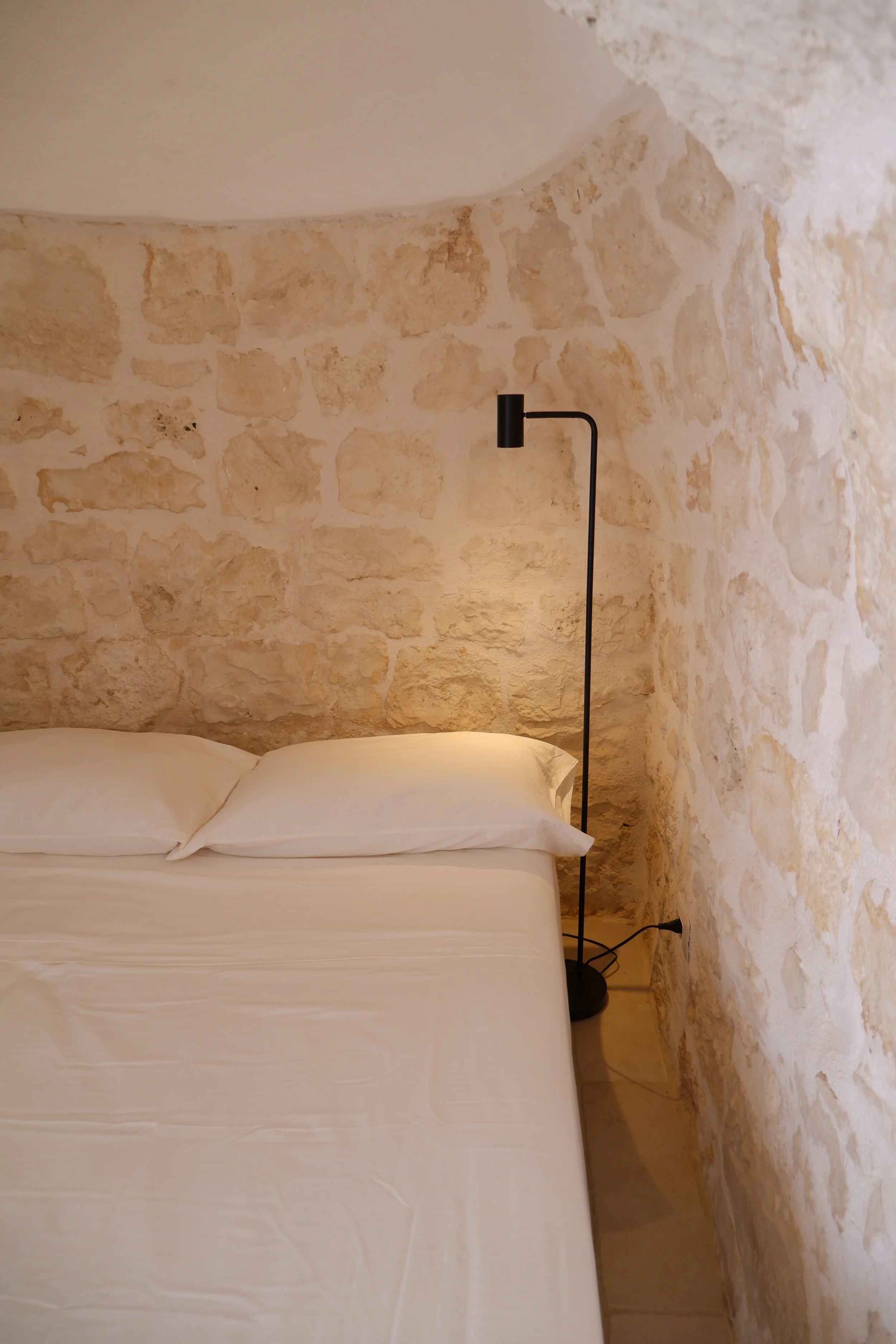 Een bed met witte lakens en twee kussens, een zwarte vloerlamp en een stenen muur in de achtergrond.