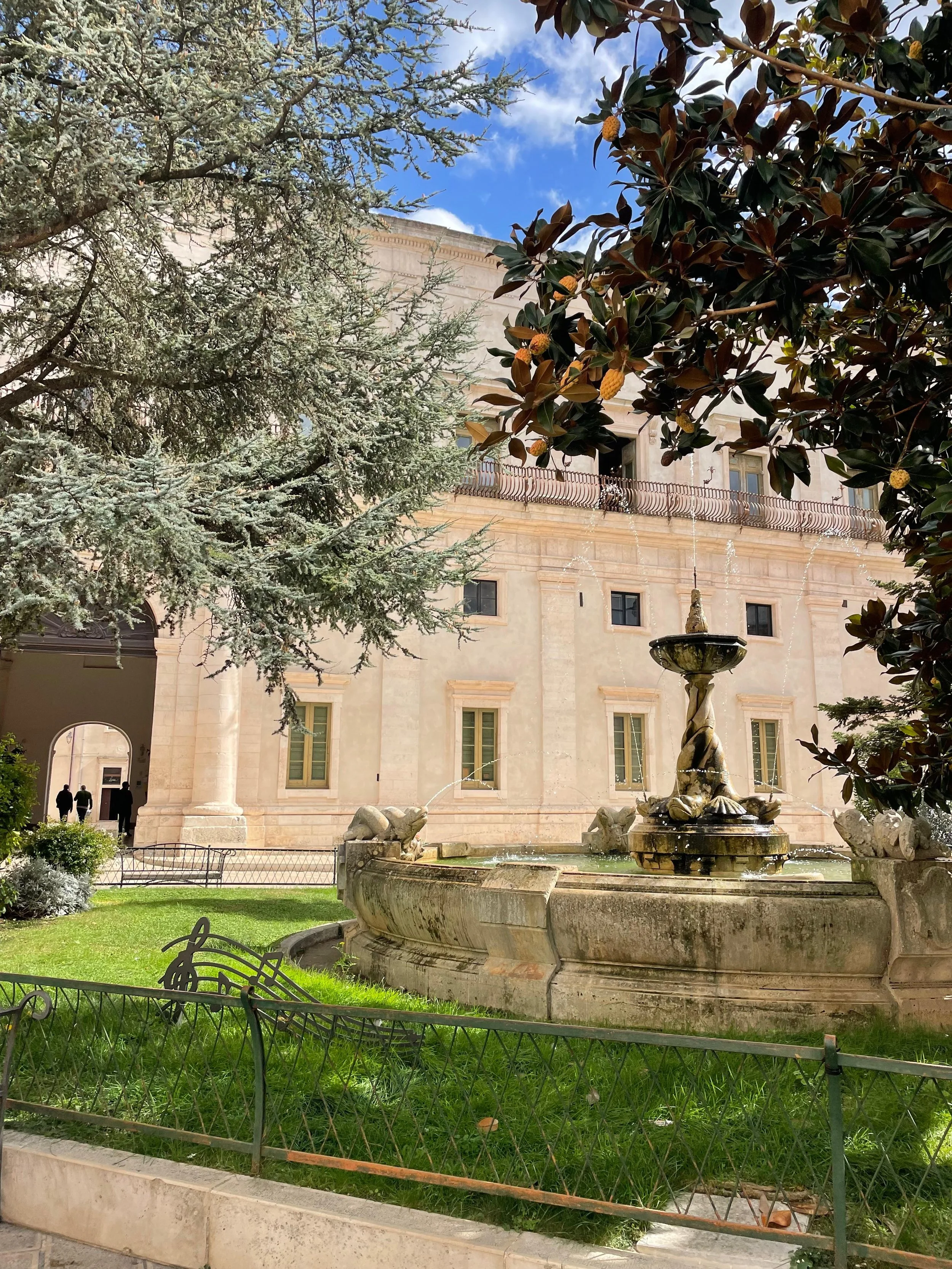 Een sierlijke fontein met beelden van vissen, omringd door groene gras en bomen, met een oude gebouw op de achtergrond en een heldere blauwe lucht.