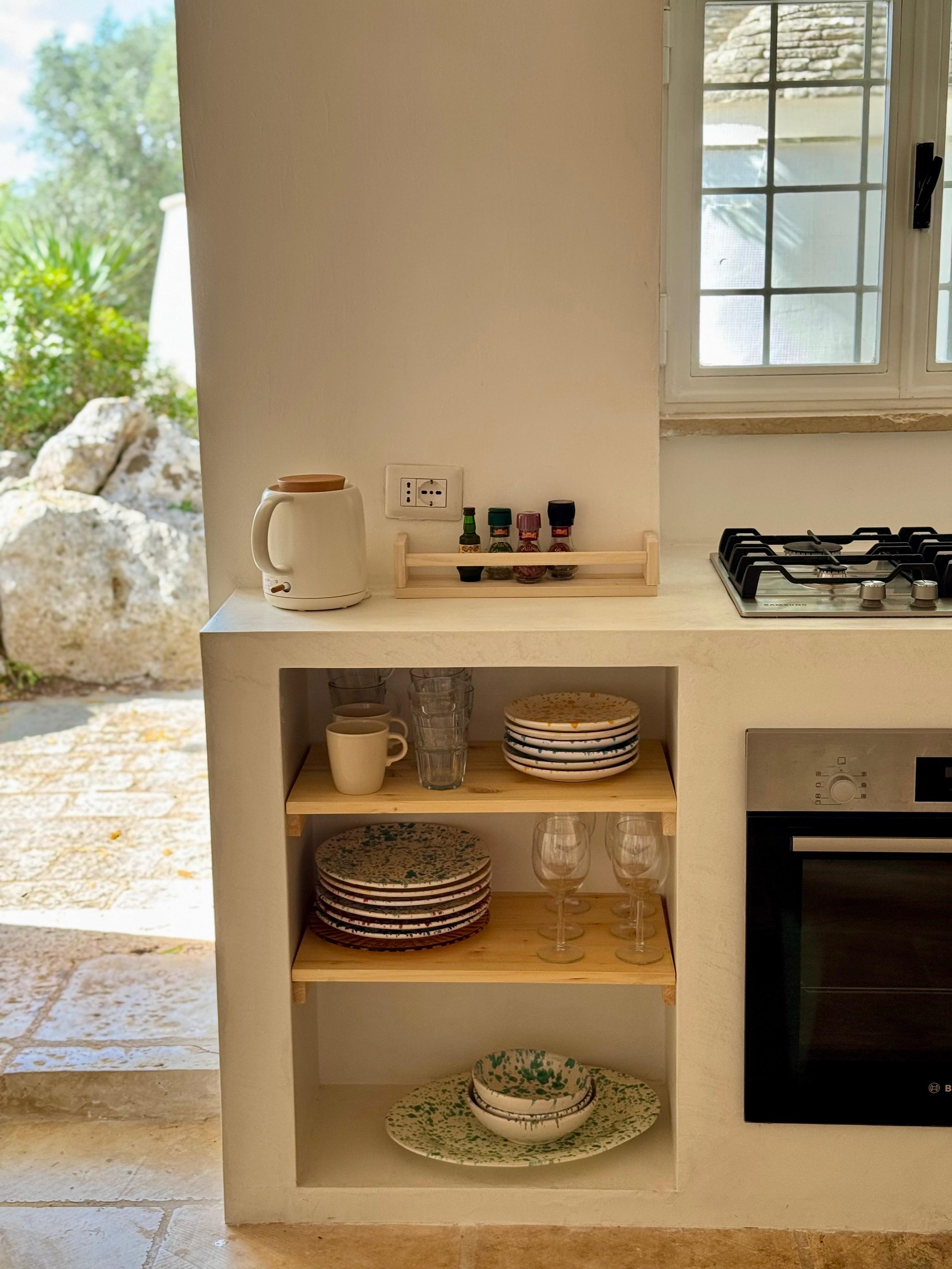 Kleine keuken met open schappen met diversa borden, glaswerk en een elektrische waterkoker, keukenraam met uitzicht op tuin en stenen pad.