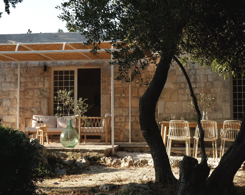 Een rustige zithoek met een bank en stoelen op een patio, omgeven door bomen en een stenen muur bij zonsondergang.