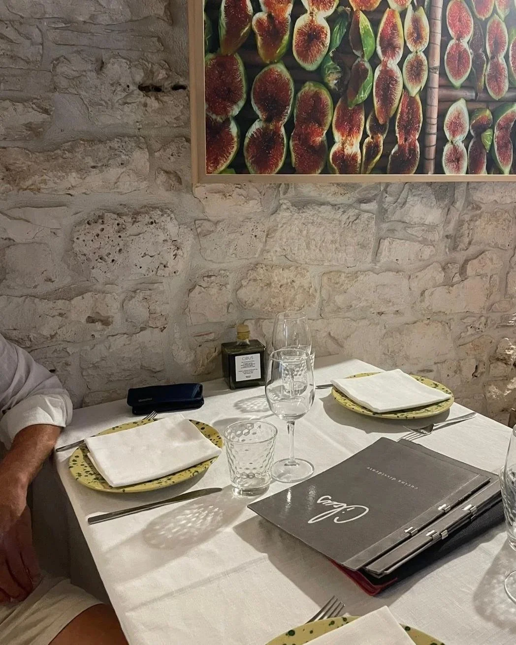 Gedeelte van een restauranttafel met placemats, bestek en glazen, tegen een bakstenen muur met een kunstwerk van vijgen.