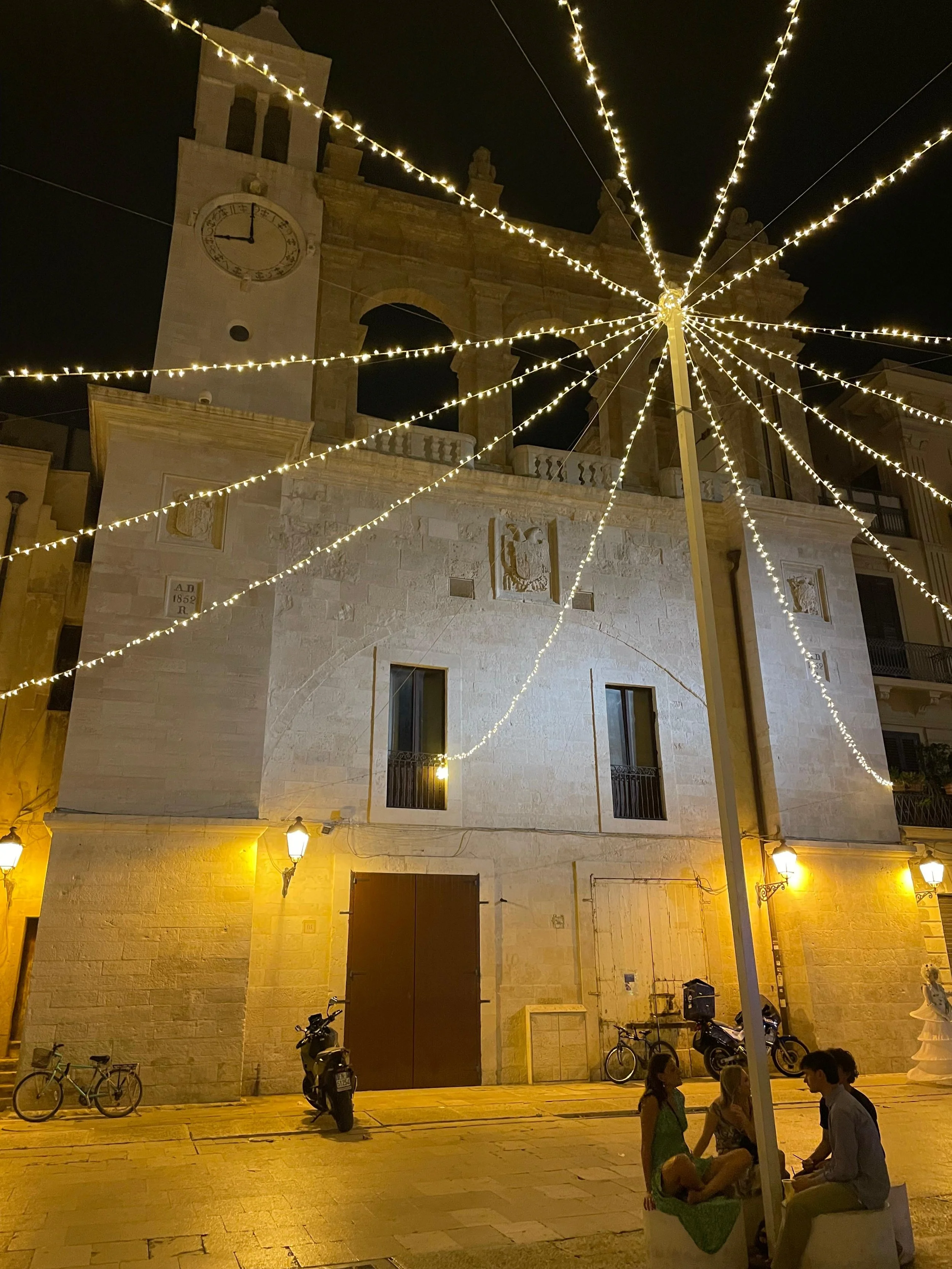 Drie mensen zitten op een bankje onder een lantaarn in een verlichte stadskade bij een oude kerk met verlichting en straatlantaarns, 's avonds.
