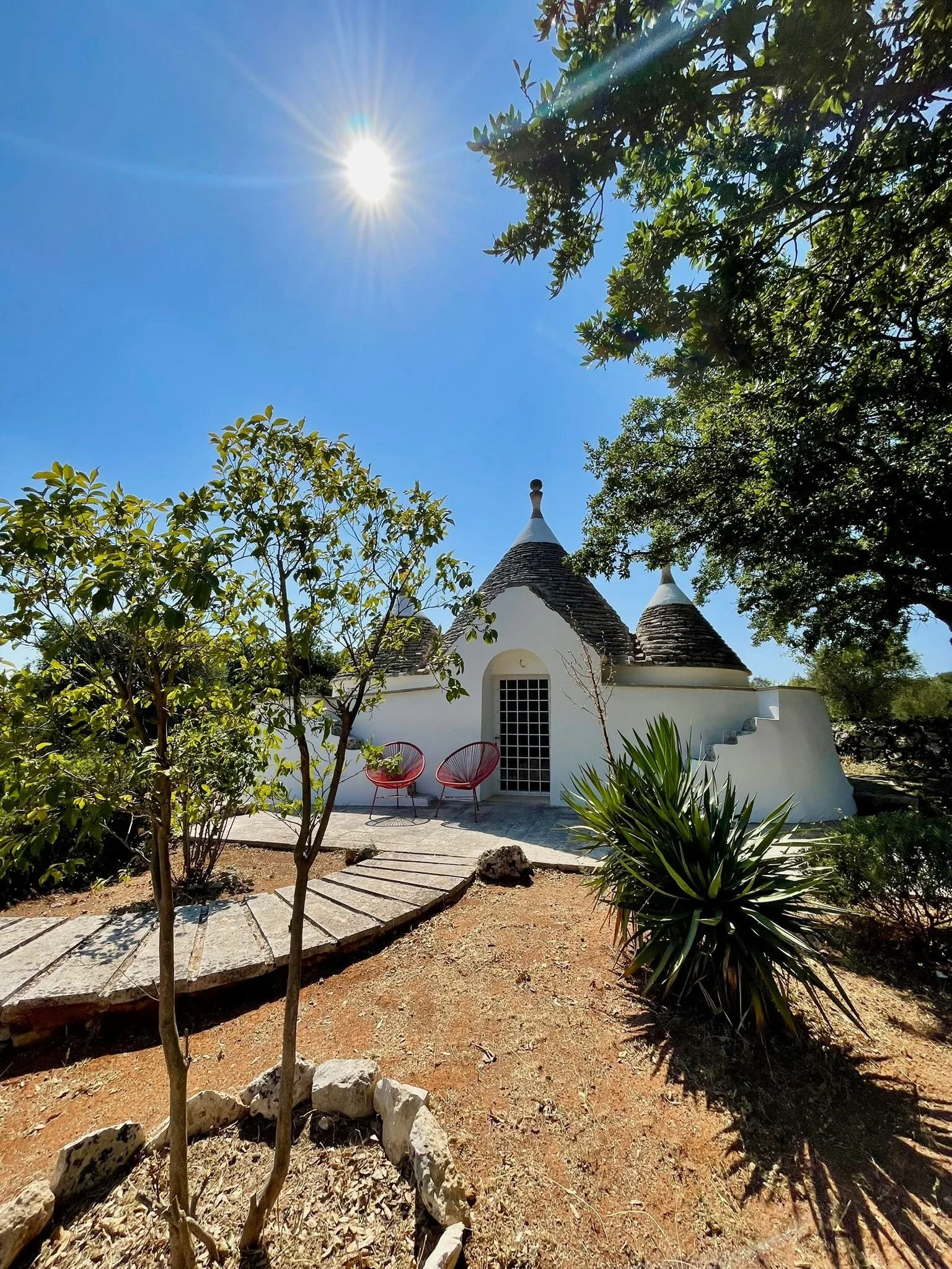 Een klein witte huisje met torenspitsen, omringd door groene bomen en een tuin op een zonnige dag.
