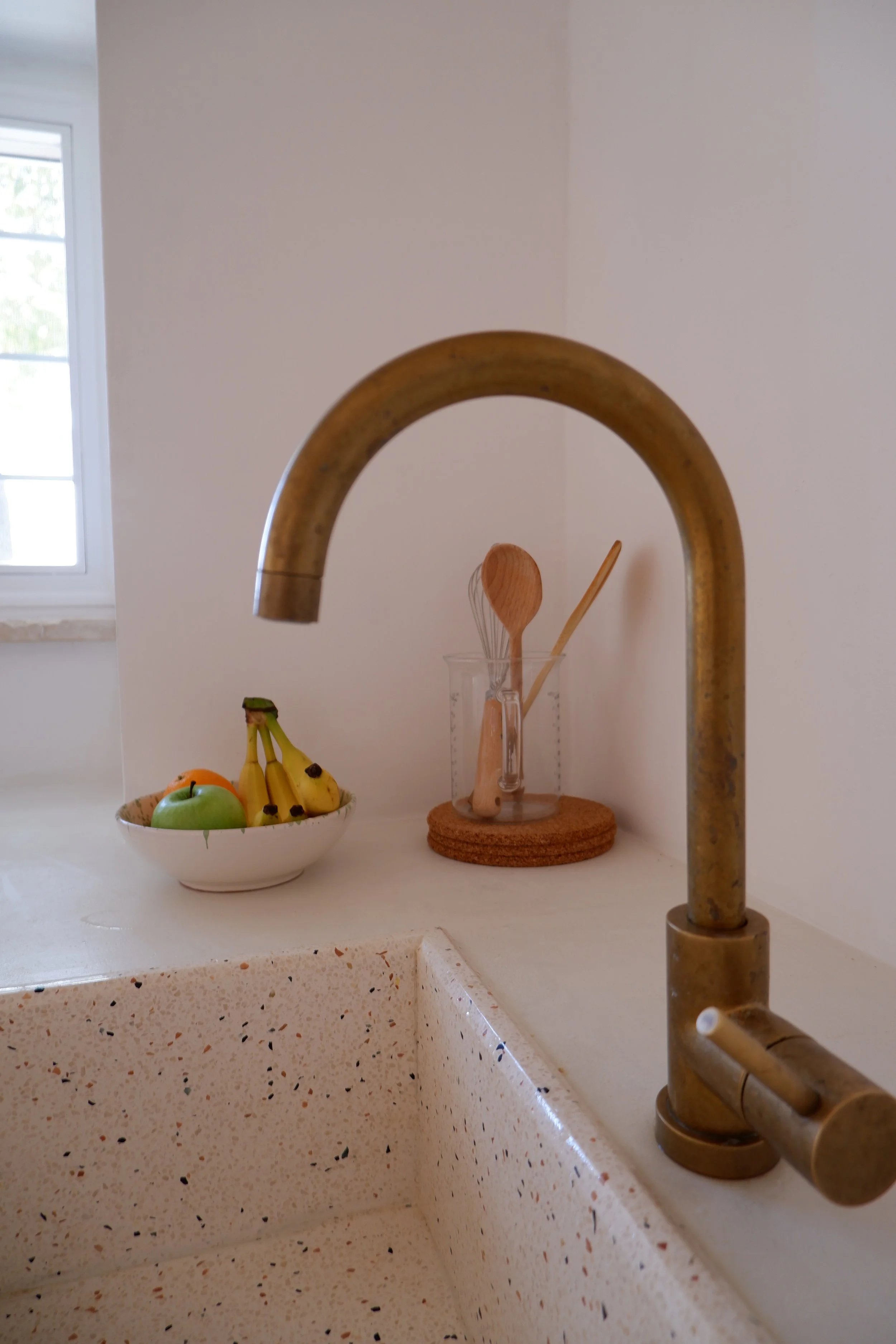 Een houten keukenmengkraan boven een gootsteen met een speckled oppervlakte. Op de achterwand staat een glas met keukenhulpmiddelen, en een kom met fruit (banaan, appel, perzik).