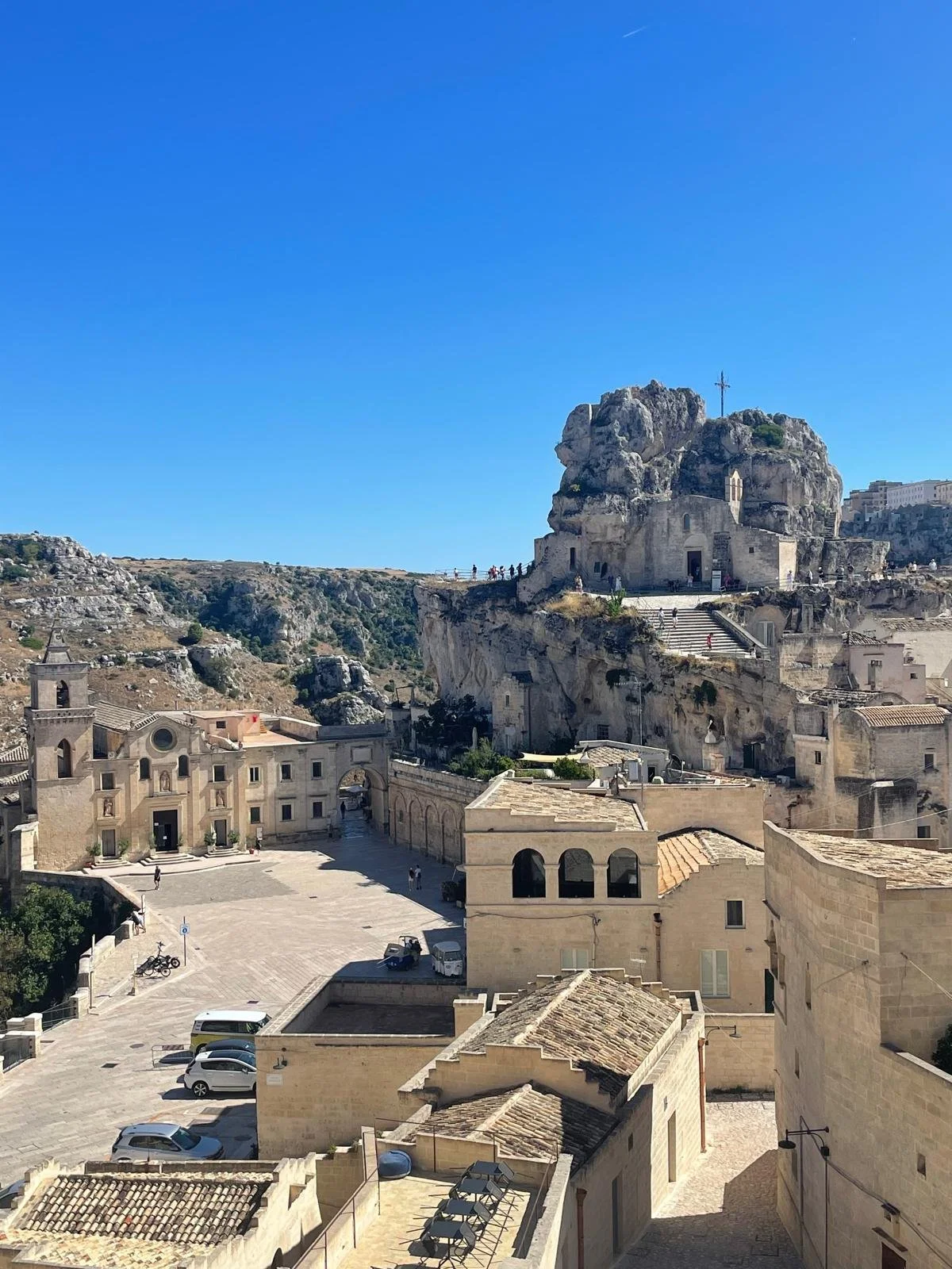 Uitzicht over een historische stad met beige gebouwen en een rotsachtig landschap onder een heldere blauwe hemel, met een kruis op de top van de rots.