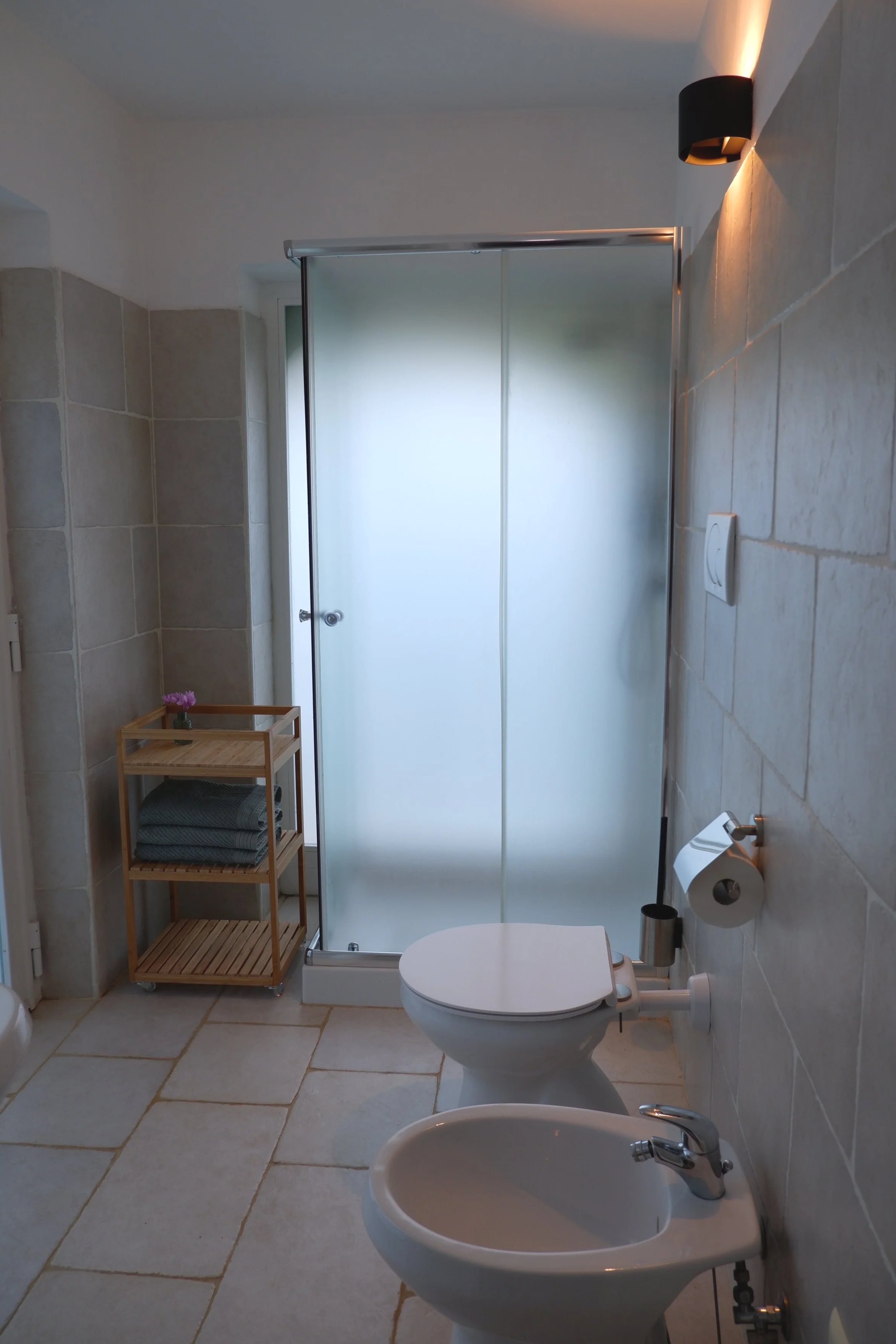 Een badkamer met een douchecabine, een toilet, een bidet, een toiletrol, een toiletborstel, een houten opbergrek met handdoeken en een vaas met bloemen.