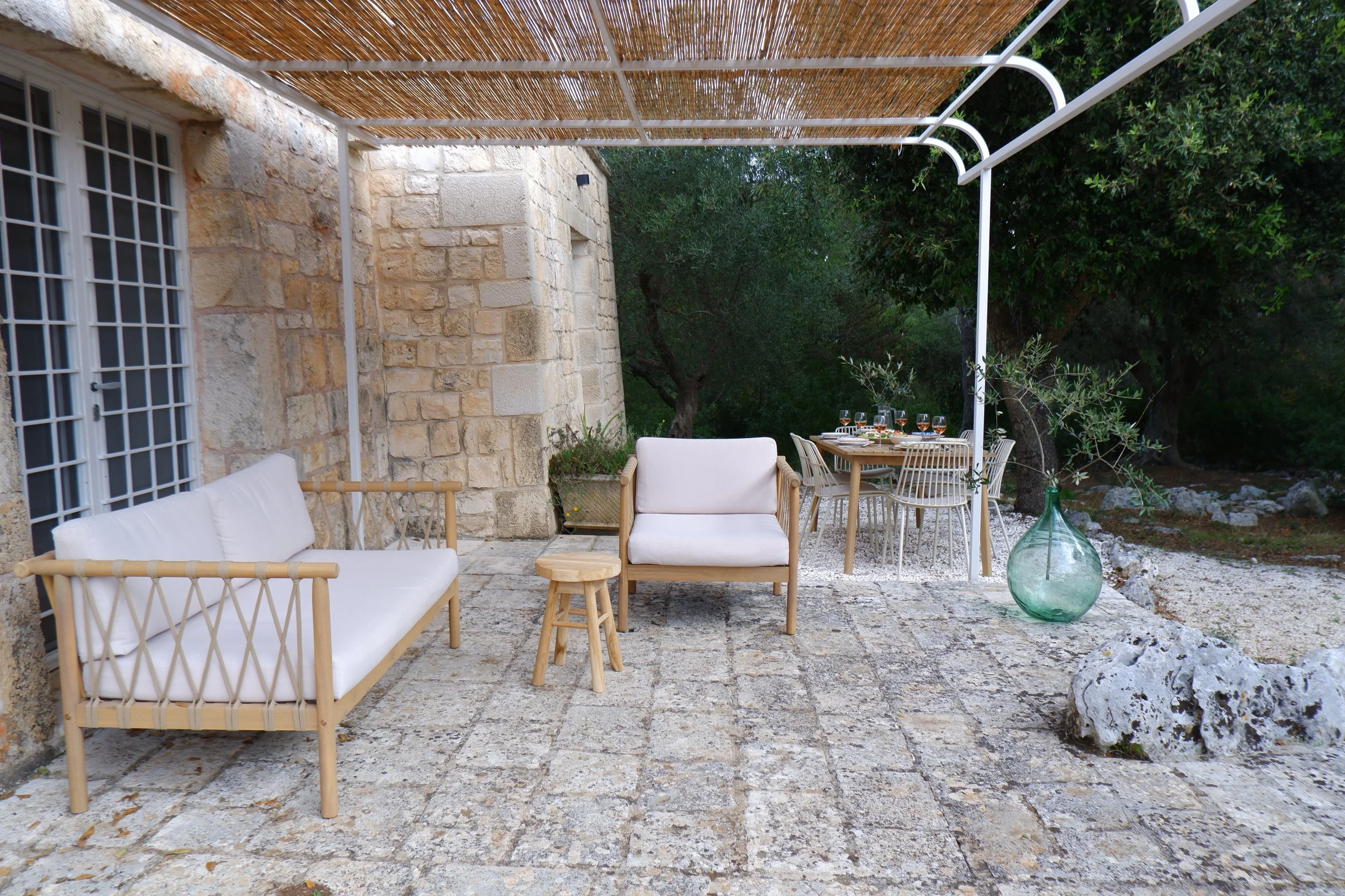 Een beschutte patio met buitenbank, stoel, kleine kruk en een tafel met eetgerei en glazen, omgeven door groene bomen en decoratieve stenen