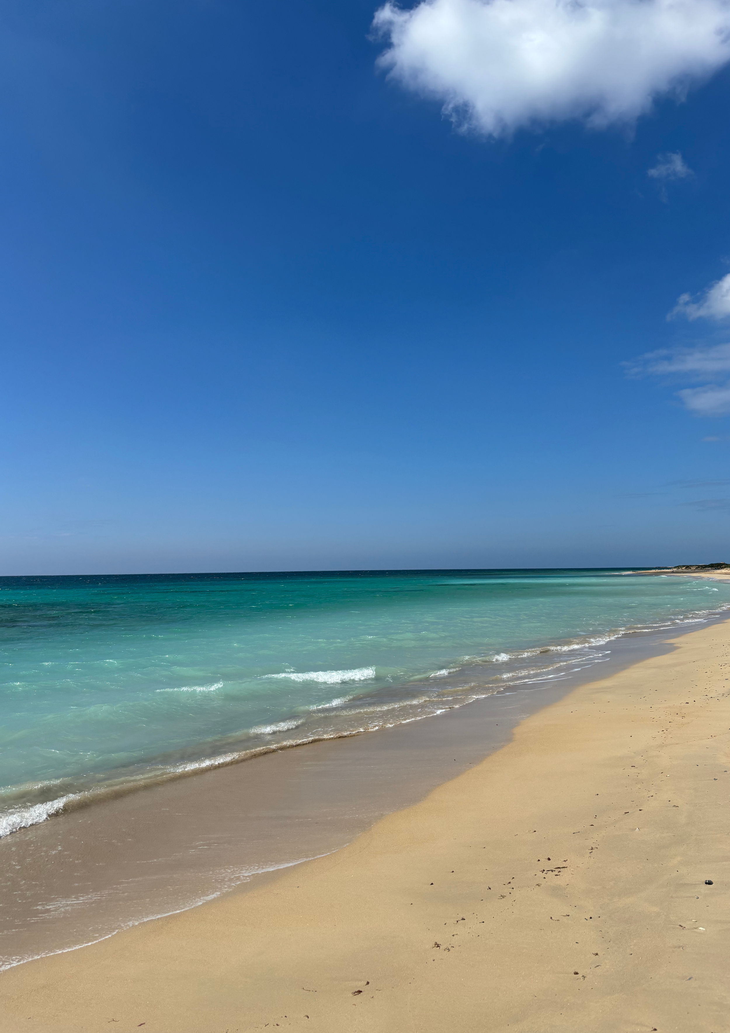 Een rustige, zonnige strand met witte zand en turquoise water onder een blauwe lucht met enkele witte wolken.