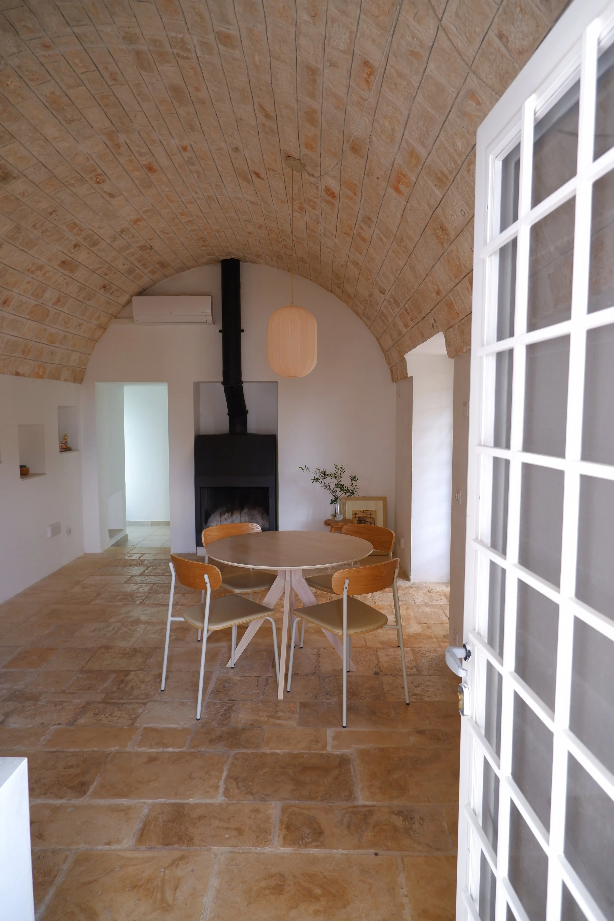 Gezellige woonkamer met een stenen boog plafond, ronde houten eettafel met vier stoelen, een moderne open haard, een hangende lamp, en een deur met vierkant raamwerk in de voorgrond.