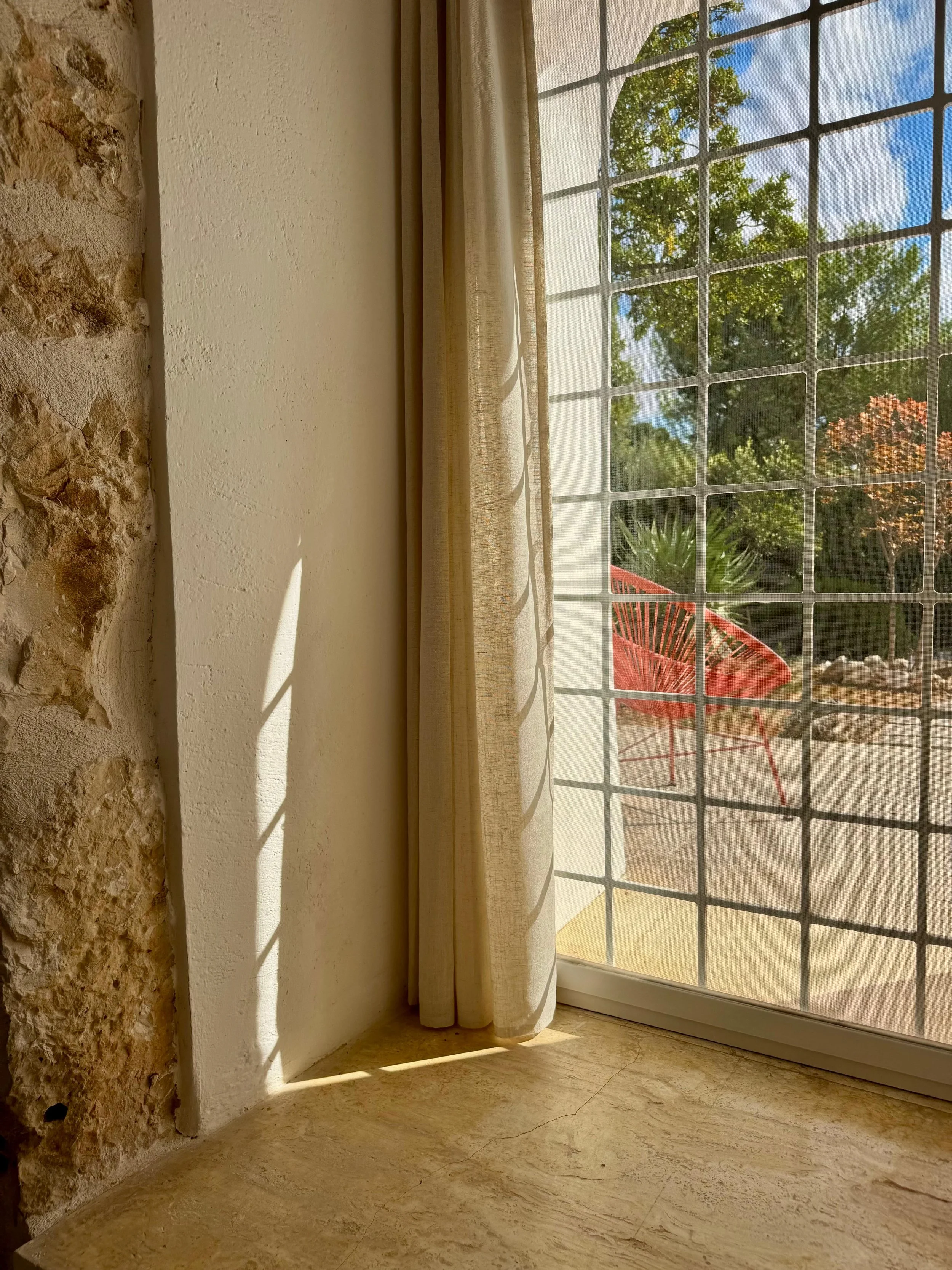 De afbeelding toont een raam met een gaasrooster, een beige gordijn en een uitzicht op een tuin met een rode stoel, bomen en een blauwe lucht.